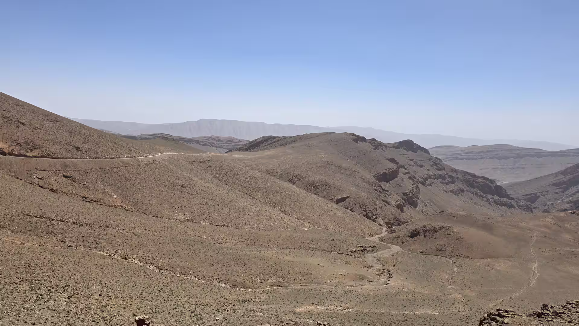 Open semi-arid mountain landscape with rolling rocky hills, a narrow road tracing the ridge, and broad canyon systems fading into the distant Atlas Mountains under a clear sky.