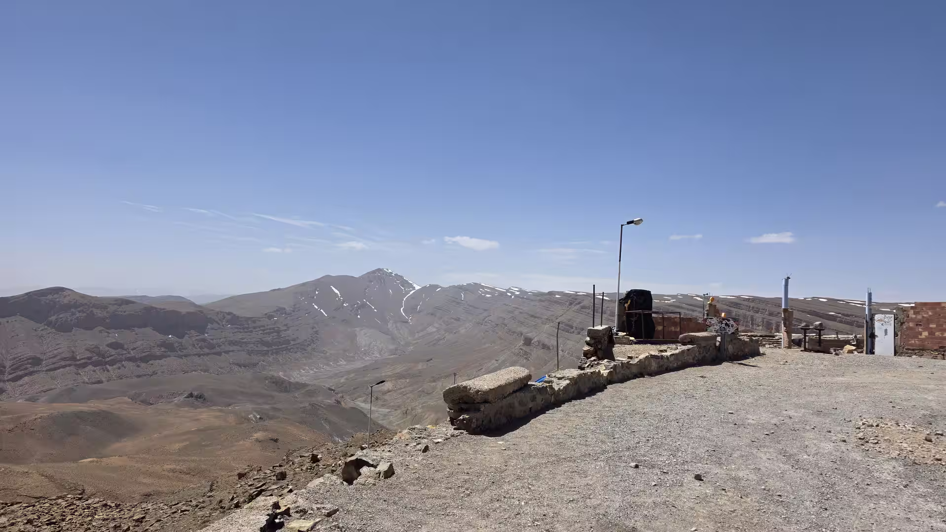 Mountain viewpoint overlooking a broad high-altitude basin and layered Atlas ridges with remaining snow patches, seen from a roadside lookout under a clear blue sky.