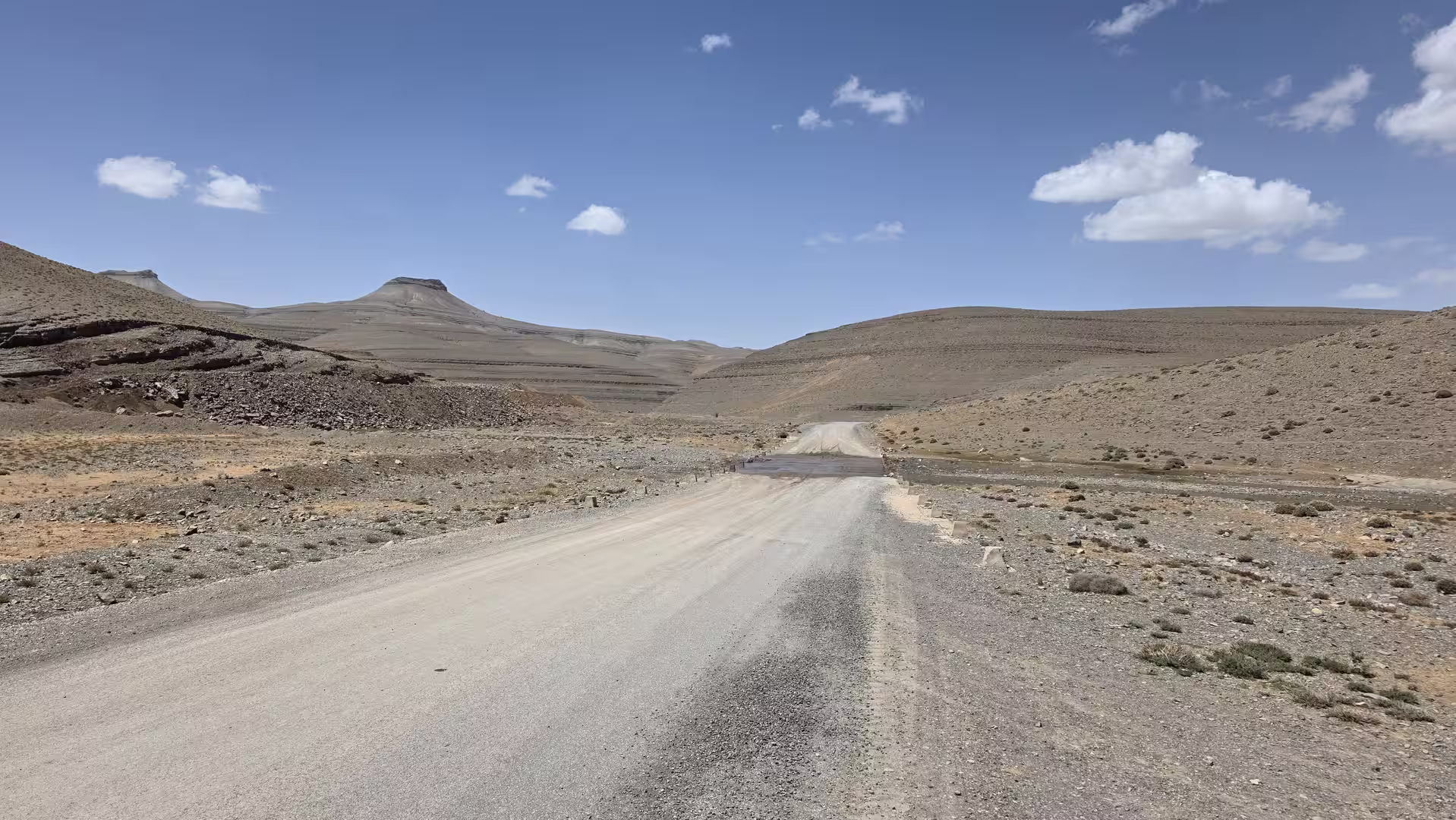 Unpaved mountain road crossing a dry high-altitude plateau with layered rock formations and isolated flat-topped hills under a bright blue sky.