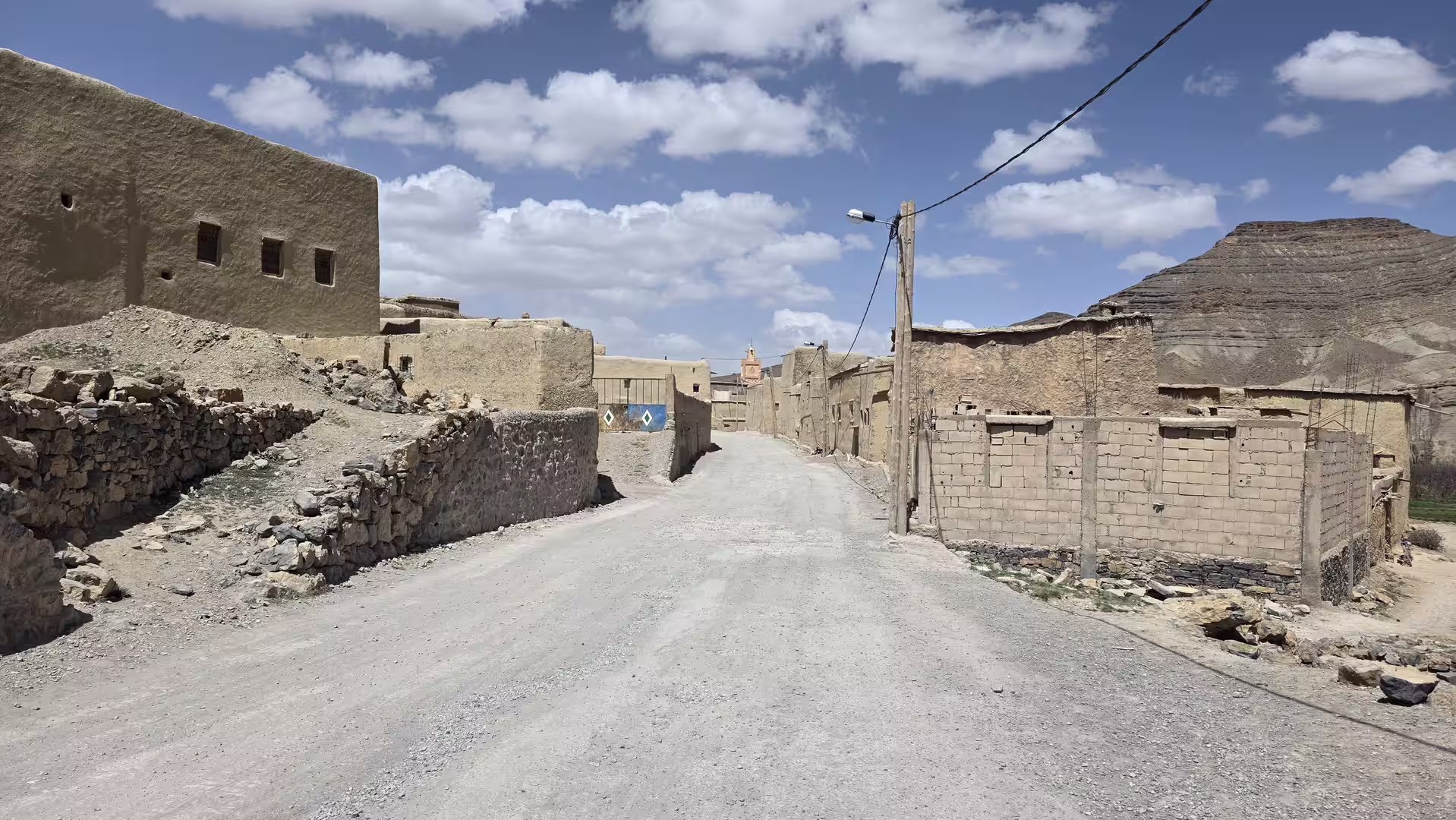Unpaved village street lined with traditional earthen houses and stone walls, set against layered hills under scattered clouds in Morocco’s mountain interior.
