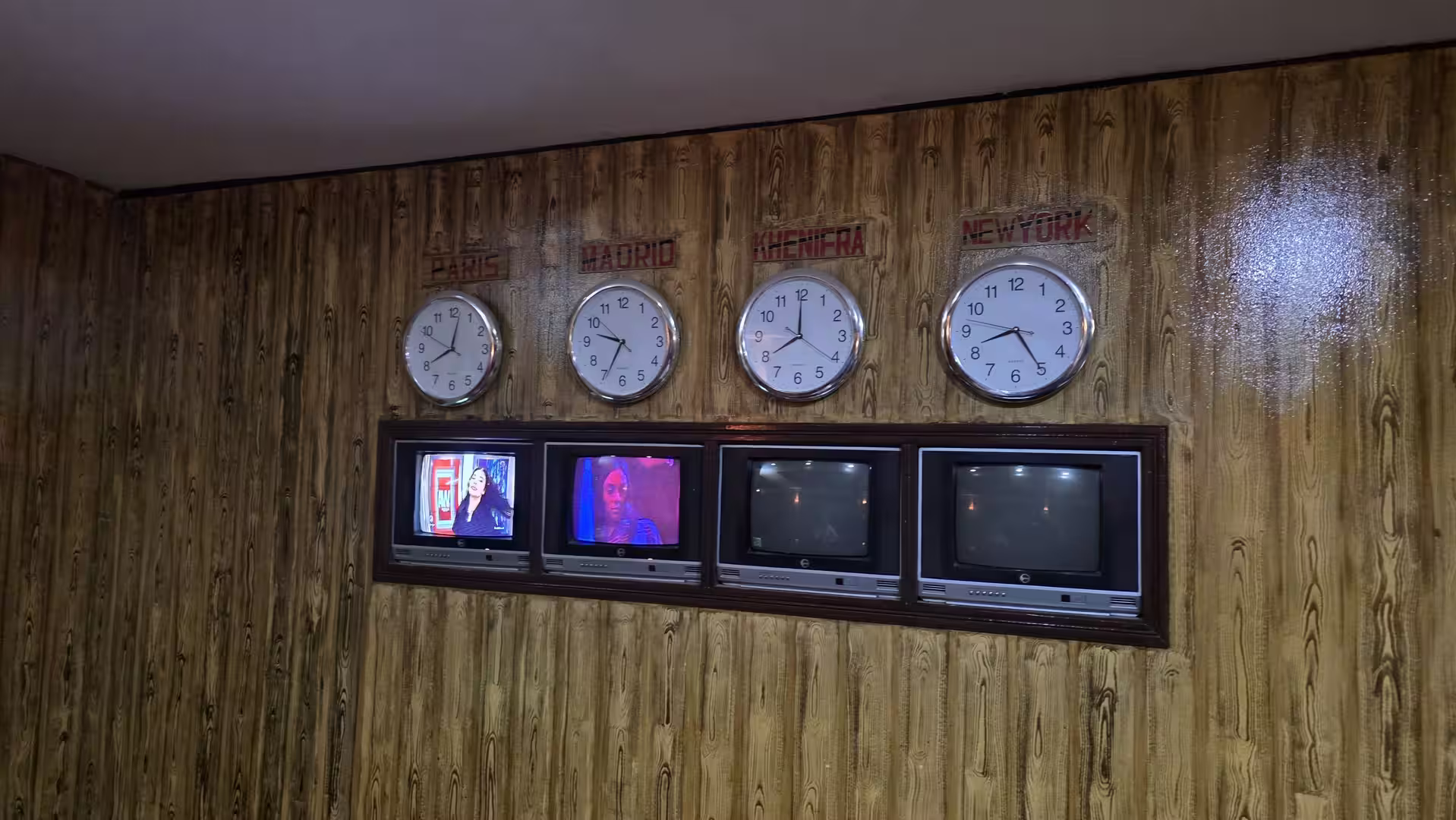 A wood-paneled wall displays four analog clocks labeled Paris, Madrid, Kinshasa, and New York above a row of vintage CRT televisions, creating a retro newsroom or broadcast-control-room atmosphere.