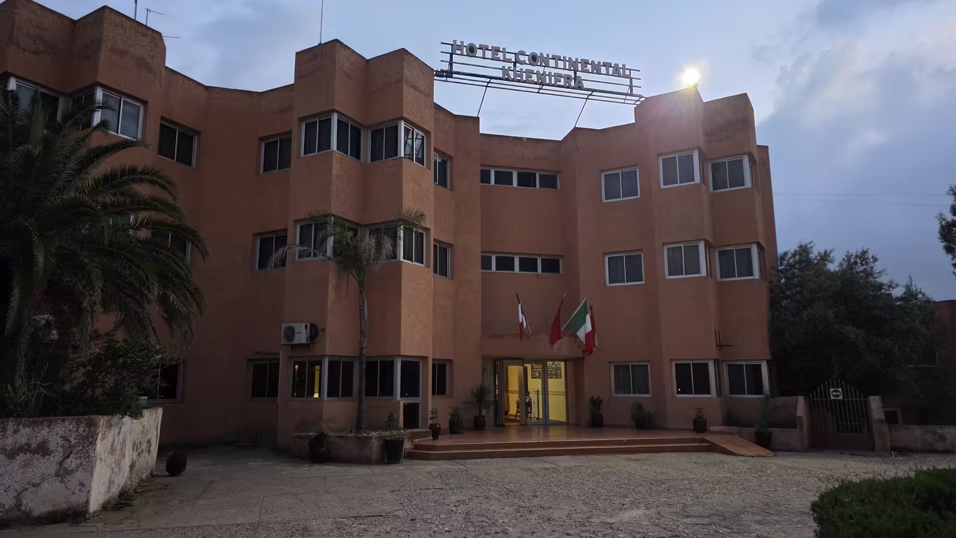 Front view of a multi-storey hotel building at dusk with a rooftop sign reading “Hotel Continental Khenifra,” a lit entrance, and several flags displayed outside.