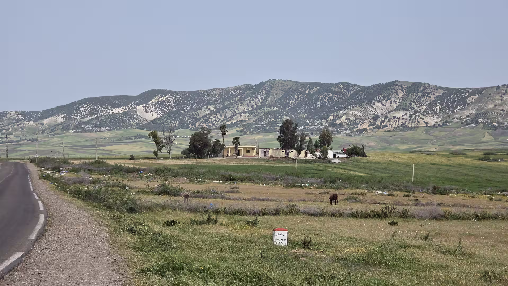 Roadside landscape near Khénifra with open fields, scattered farm buildings, and forested hills in the background.