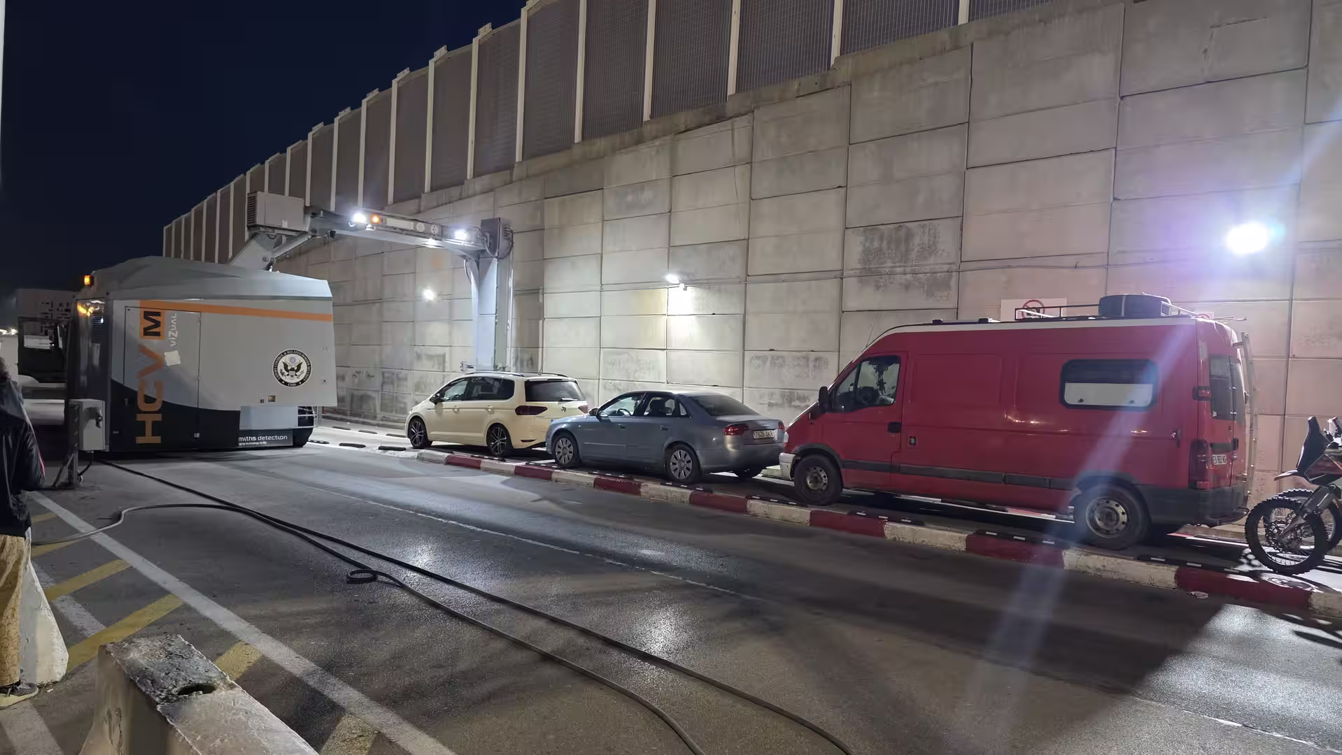 Vehicles queue at a border control checkpoint at night while a mobile cargo and vehicle inspection scanner operates beside the roadway.