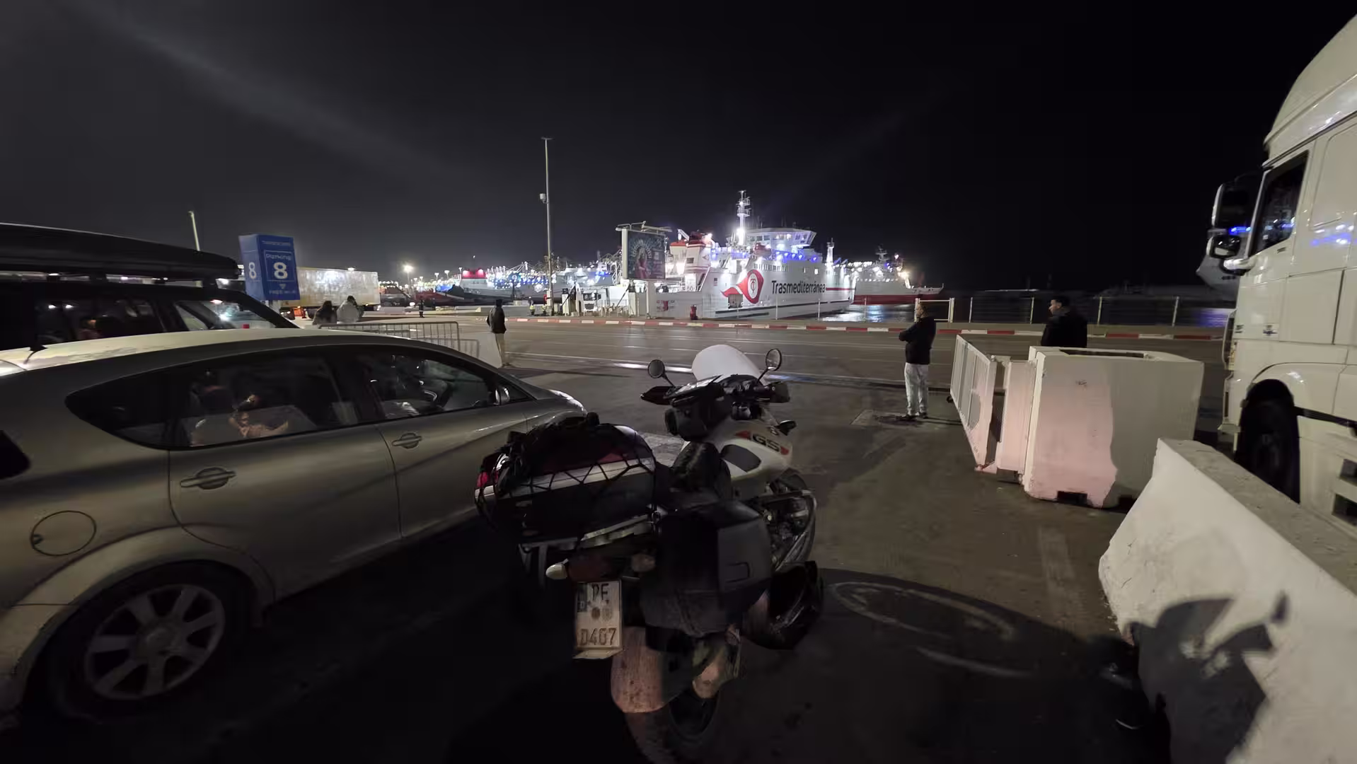 Nighttime scene at the Tanger Med ferry terminal with parked vehicles, a touring motorcycle, and an illuminated ferry preparing for passenger and vehicle boarding.