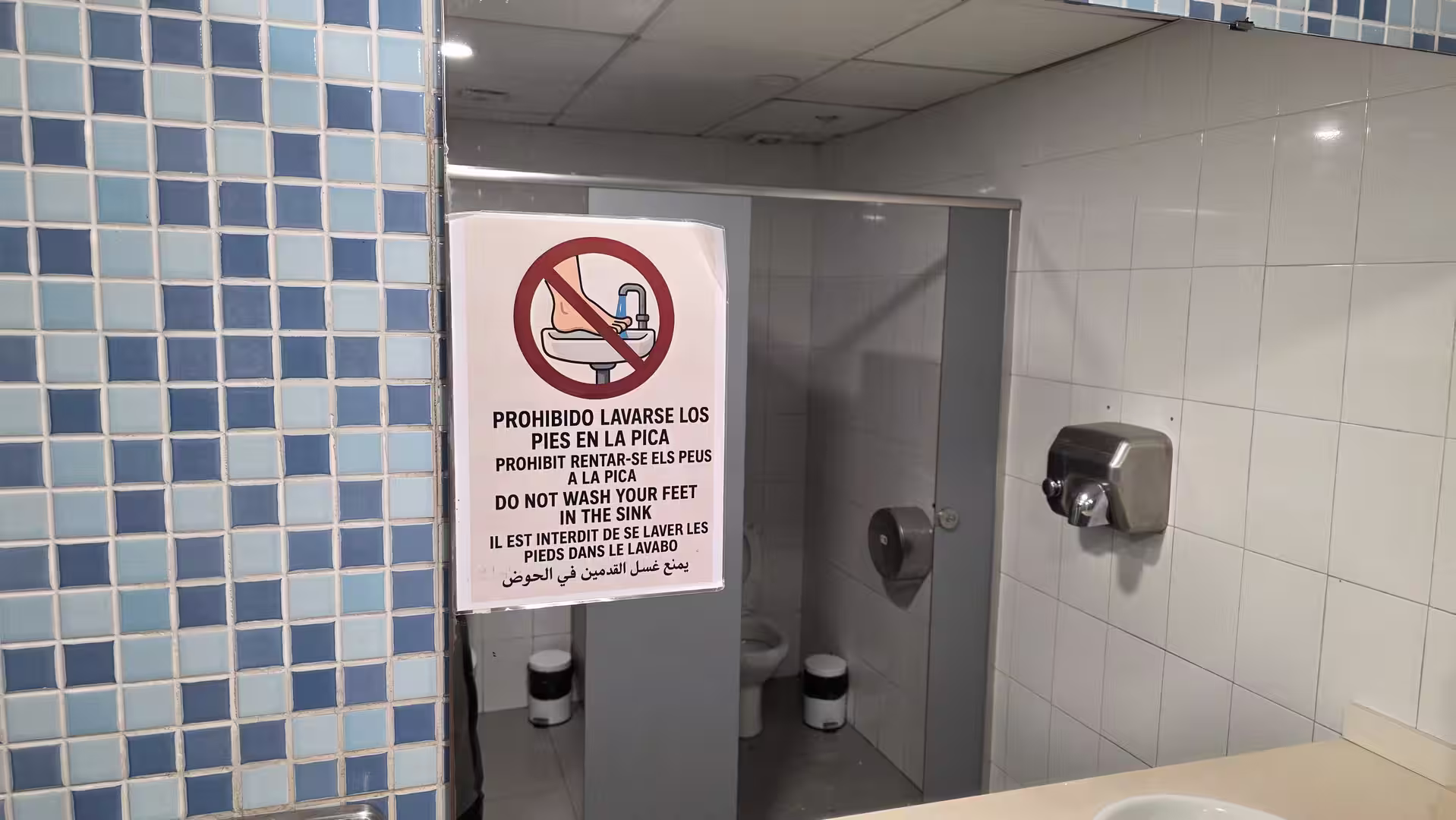 Public restroom interior with tiled walls and a multilingual sign showing that washing feet in the sink is prohibited.