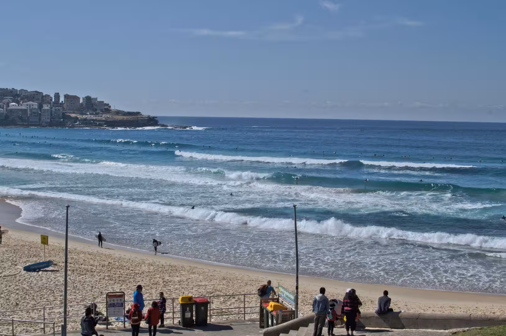 White surf breaks across turquoise water as surfers wait off a sandy beach concourse.