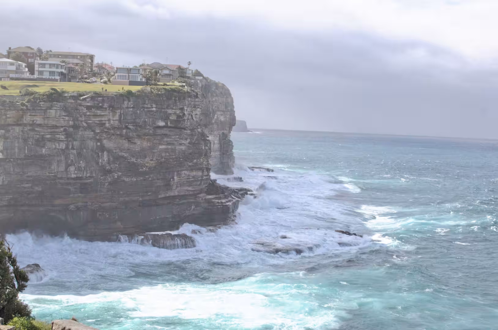Turquoise surf crashes against dark coastal cliffs with homes perched on the clifftop above.