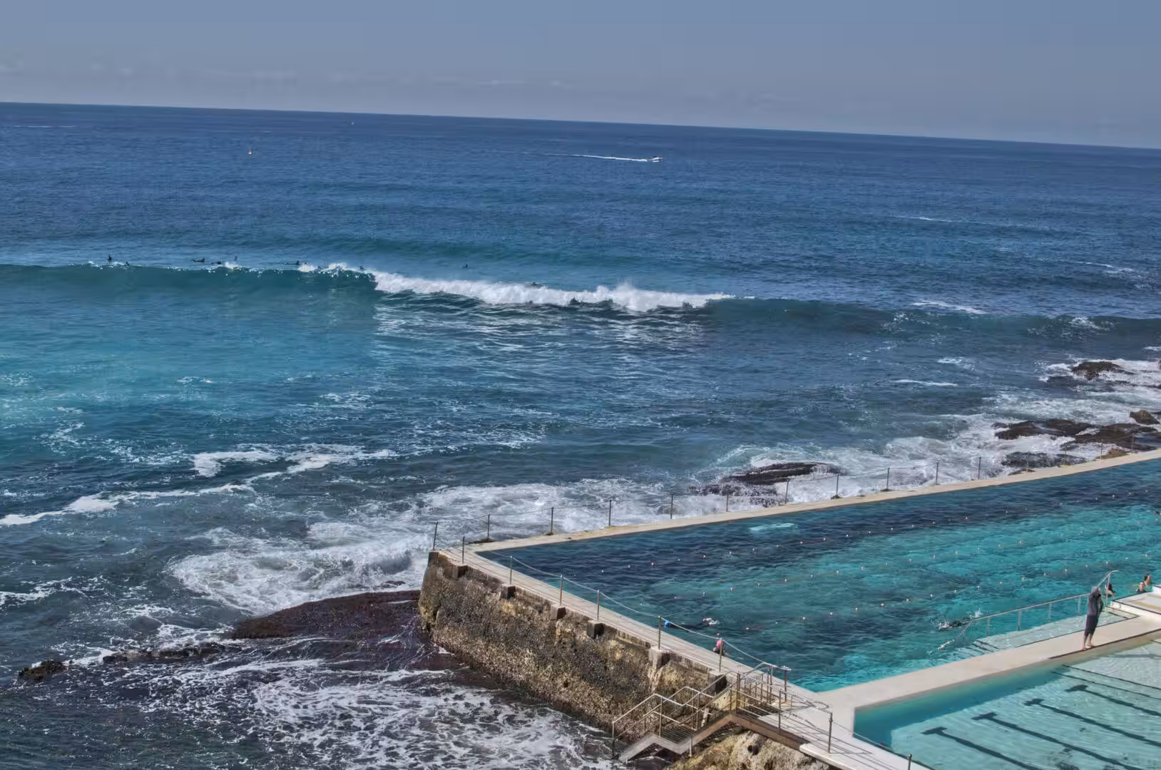 A clear saltwater pool sits in the rock while the ocean crashes against the ledge beyond.