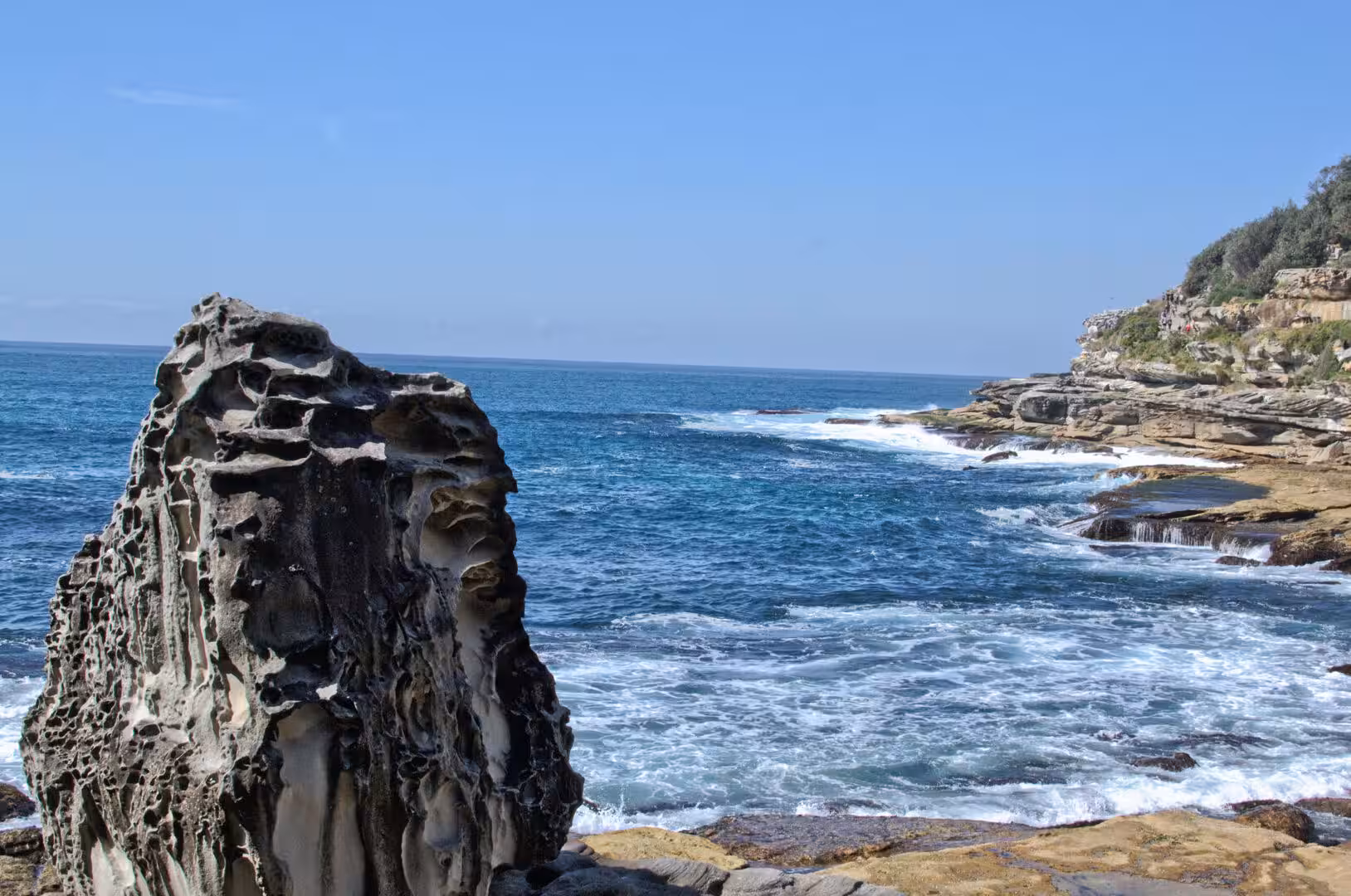 A hollowed sandstone rock stands by the deep blue ocean as surf crashes on the rocky shore.
