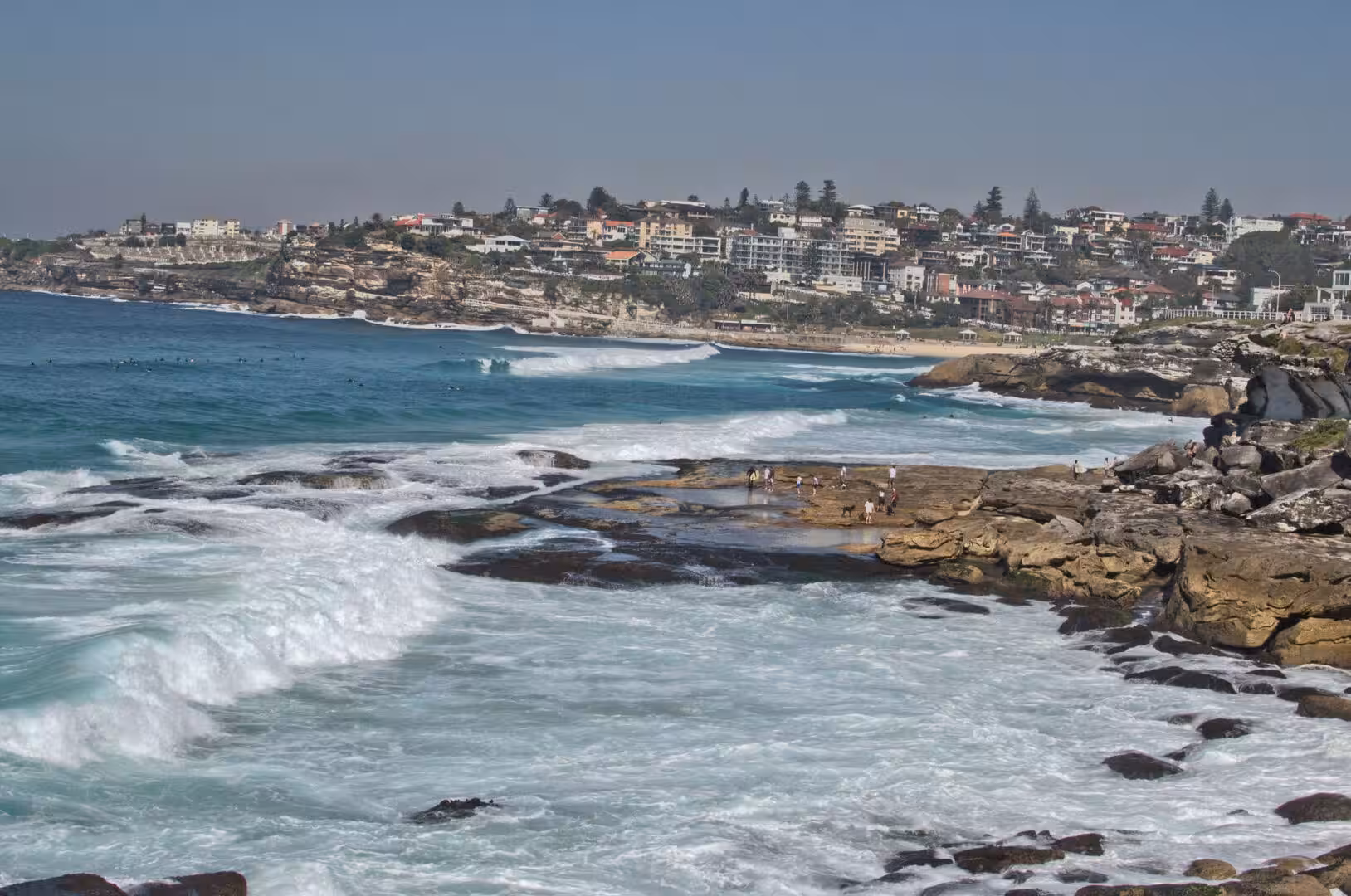 Visitors stand on the flat rocks while waves blast foam around them.