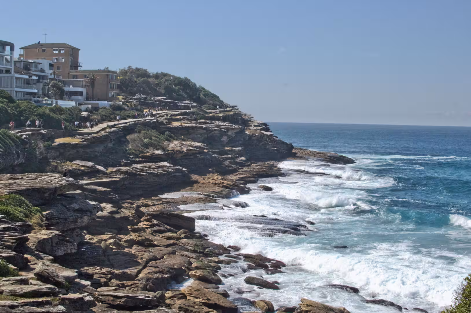 Walkers follow the path along the rocky cliff above the crashing waves.