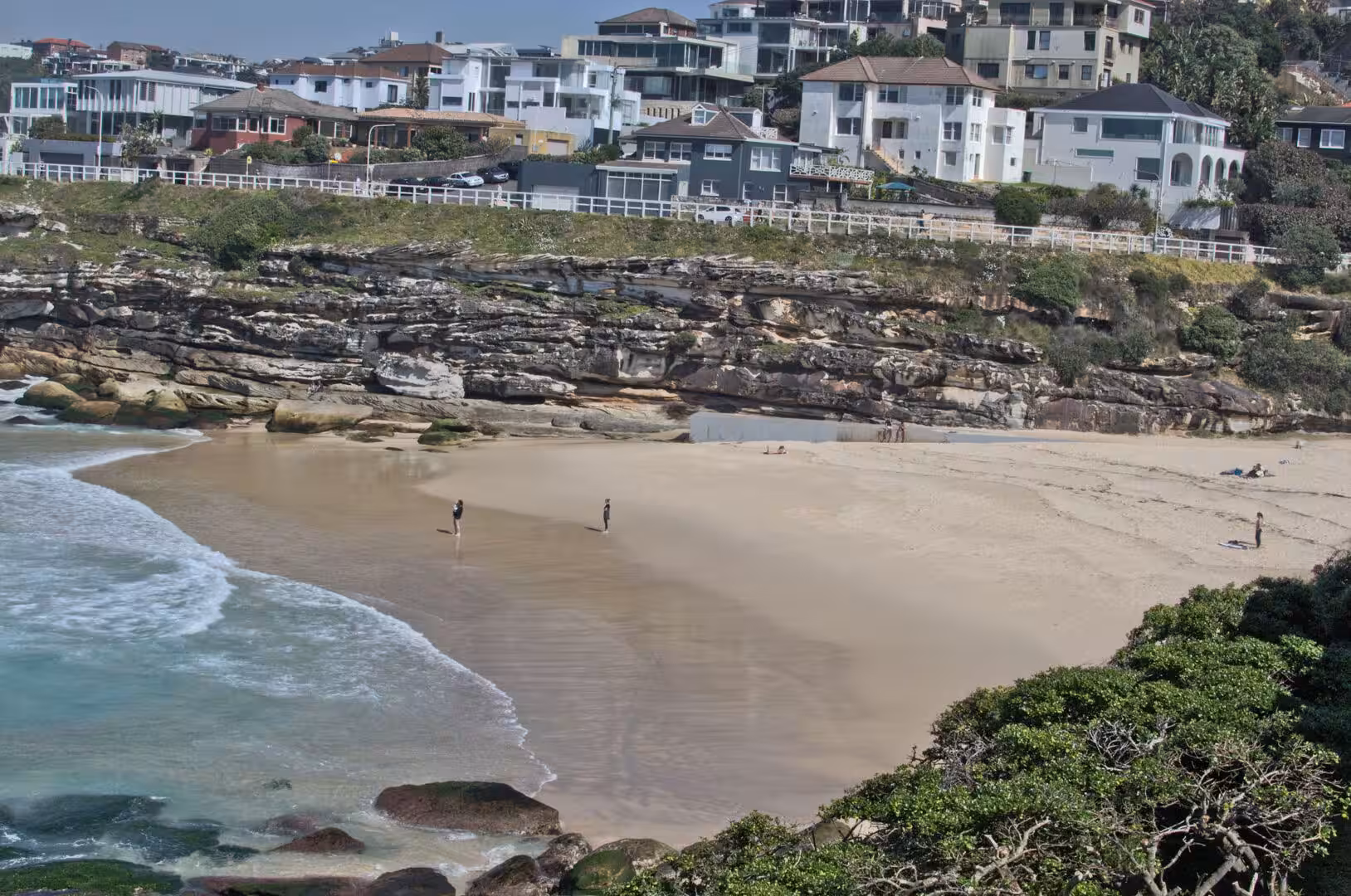 A near-empty sandy cove sits below the homes on the rocky cliff.
