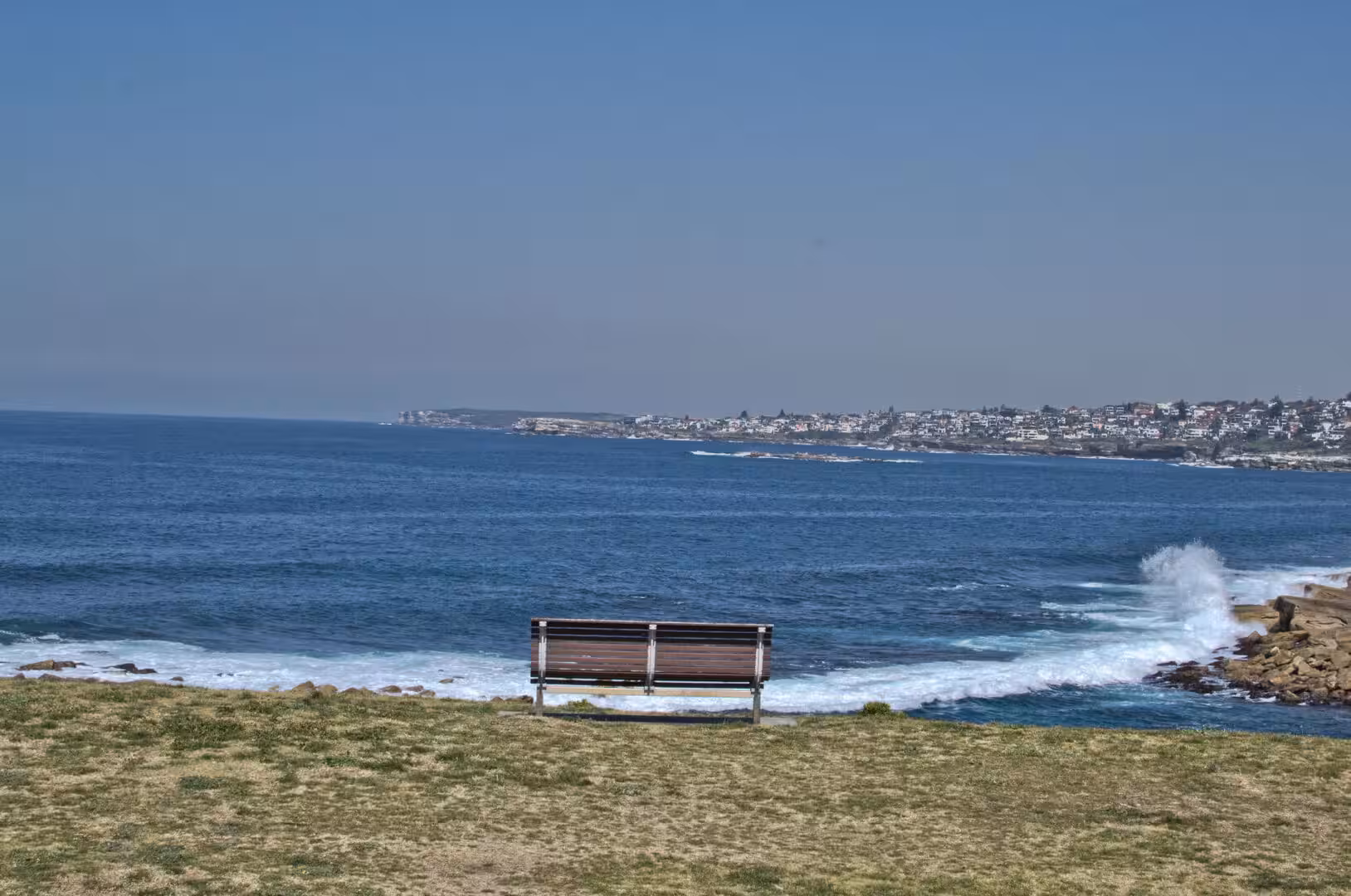 An empty bench faces the ocean from a grassy clifftop.