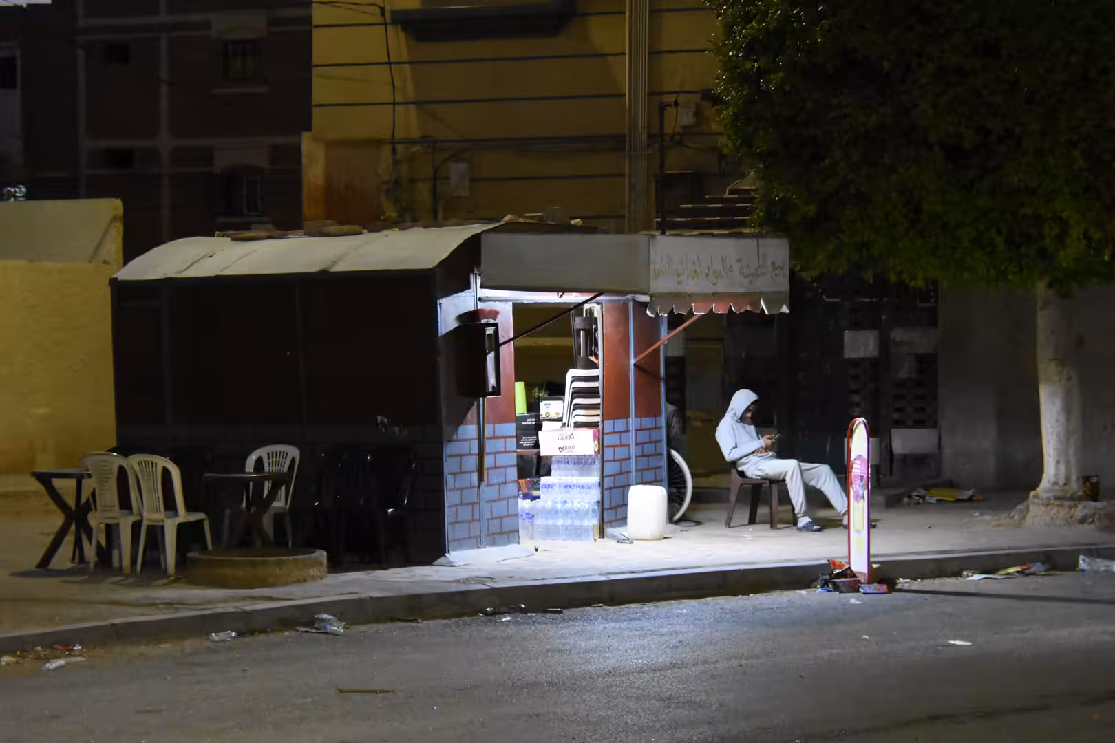 Small illuminated street café at night in Laâyoune with outdoor plastic chairs and a person sitting beside the entrance.
