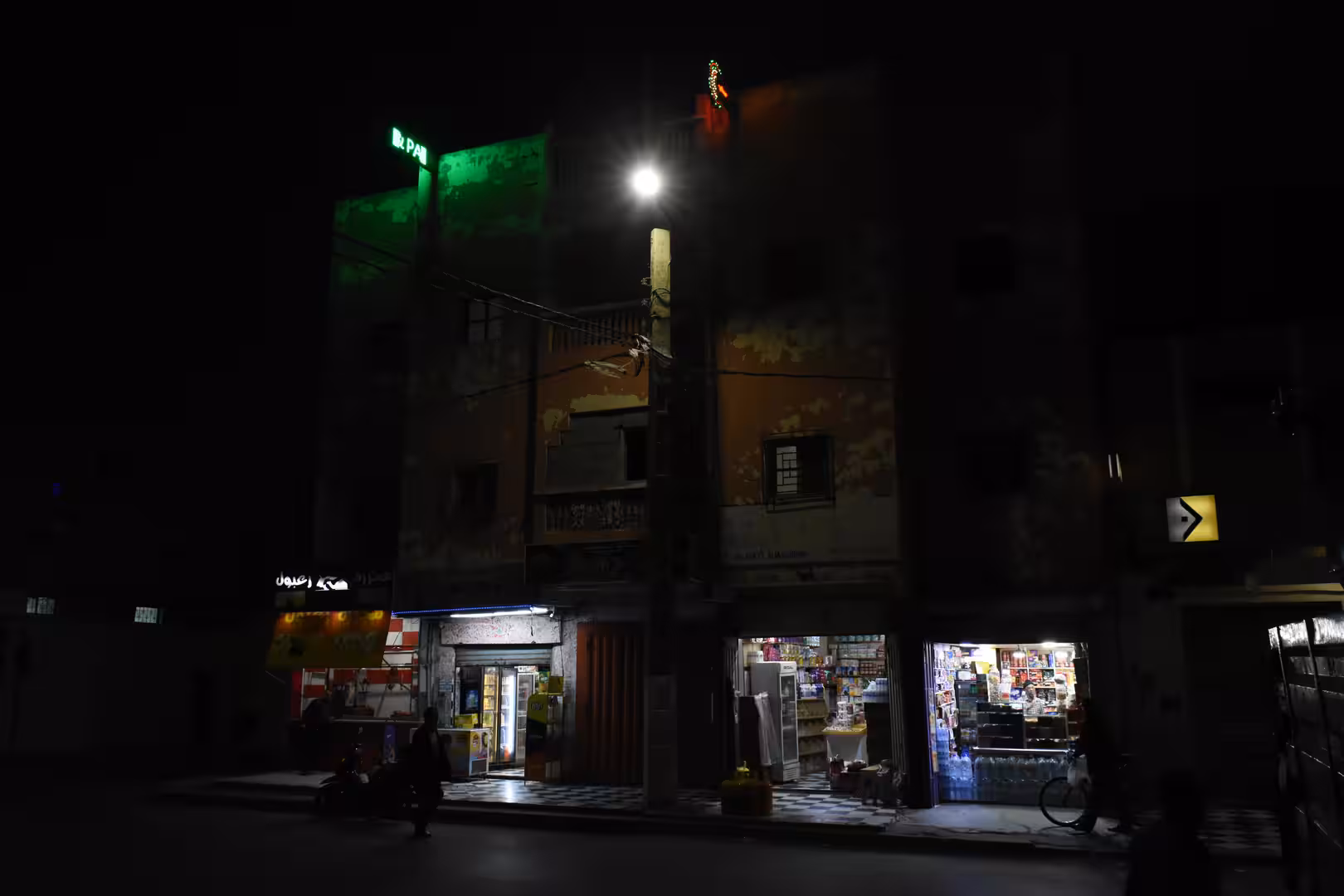 Nighttime street view in Laâyoune showing a mid-rise residential building with illuminated ground-floor shops, a streetlight and a few pedestrians.