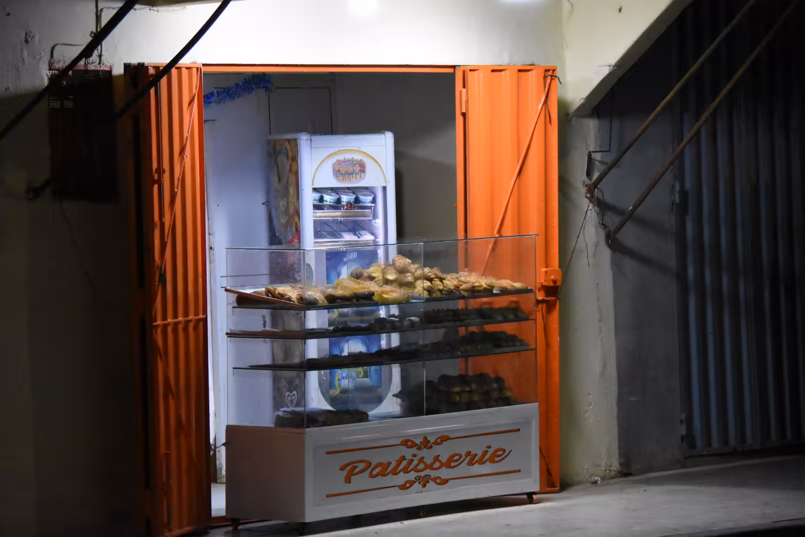 Small pastry shop open at night in Laâyoune with a glass display case of baked goods, bright interior lighting and orange metal doors.