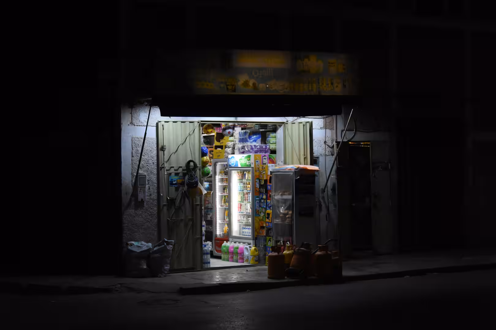 Small neighborhood convenience store in Laâyoune illuminated at night, with refrigerators, shelves of household goods and beverages visible through open metal doors.