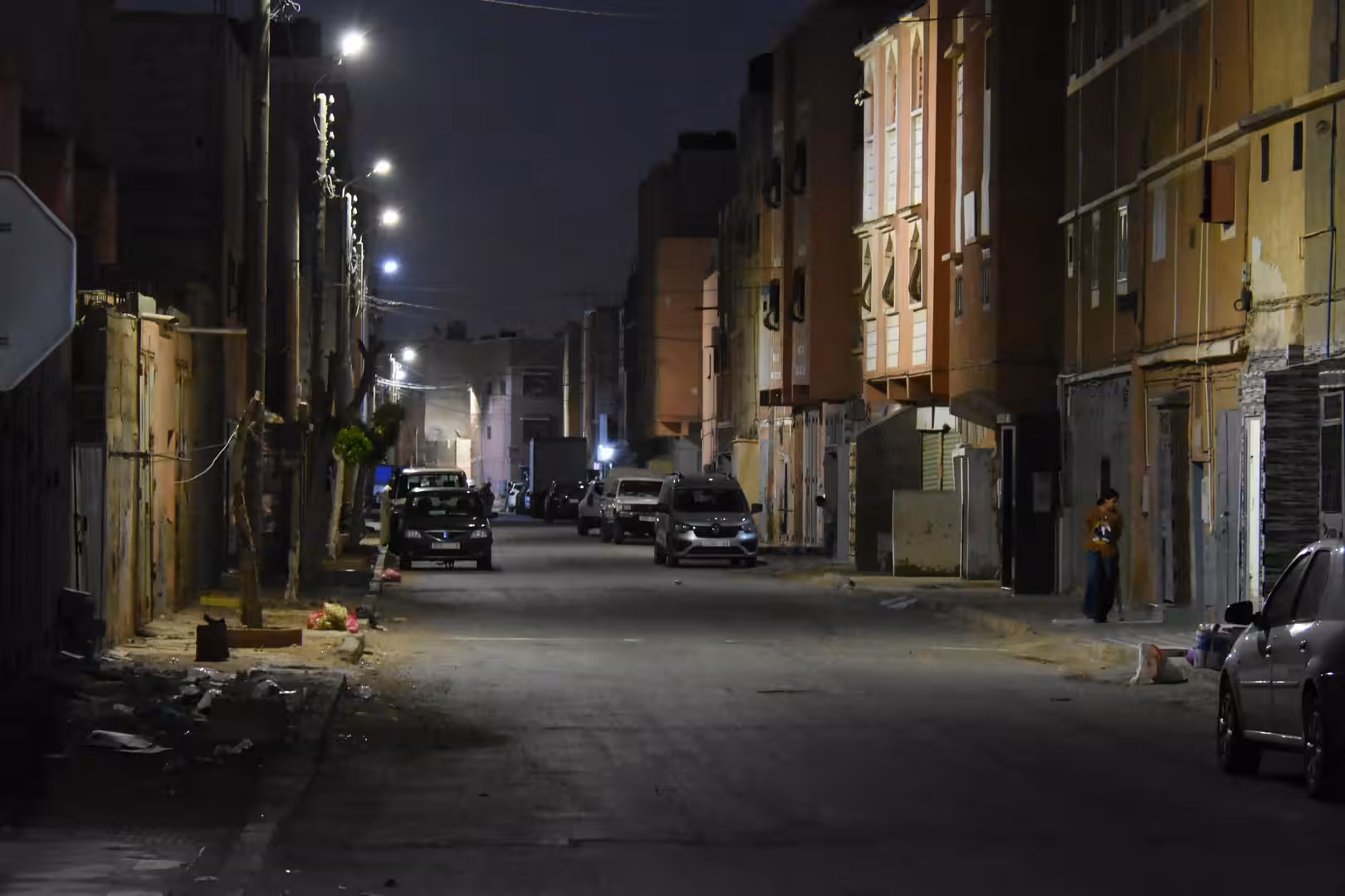 Quiet residential street in Laâyoune at night with apartment buildings, parked vehicles and streetlights extending into the distance.