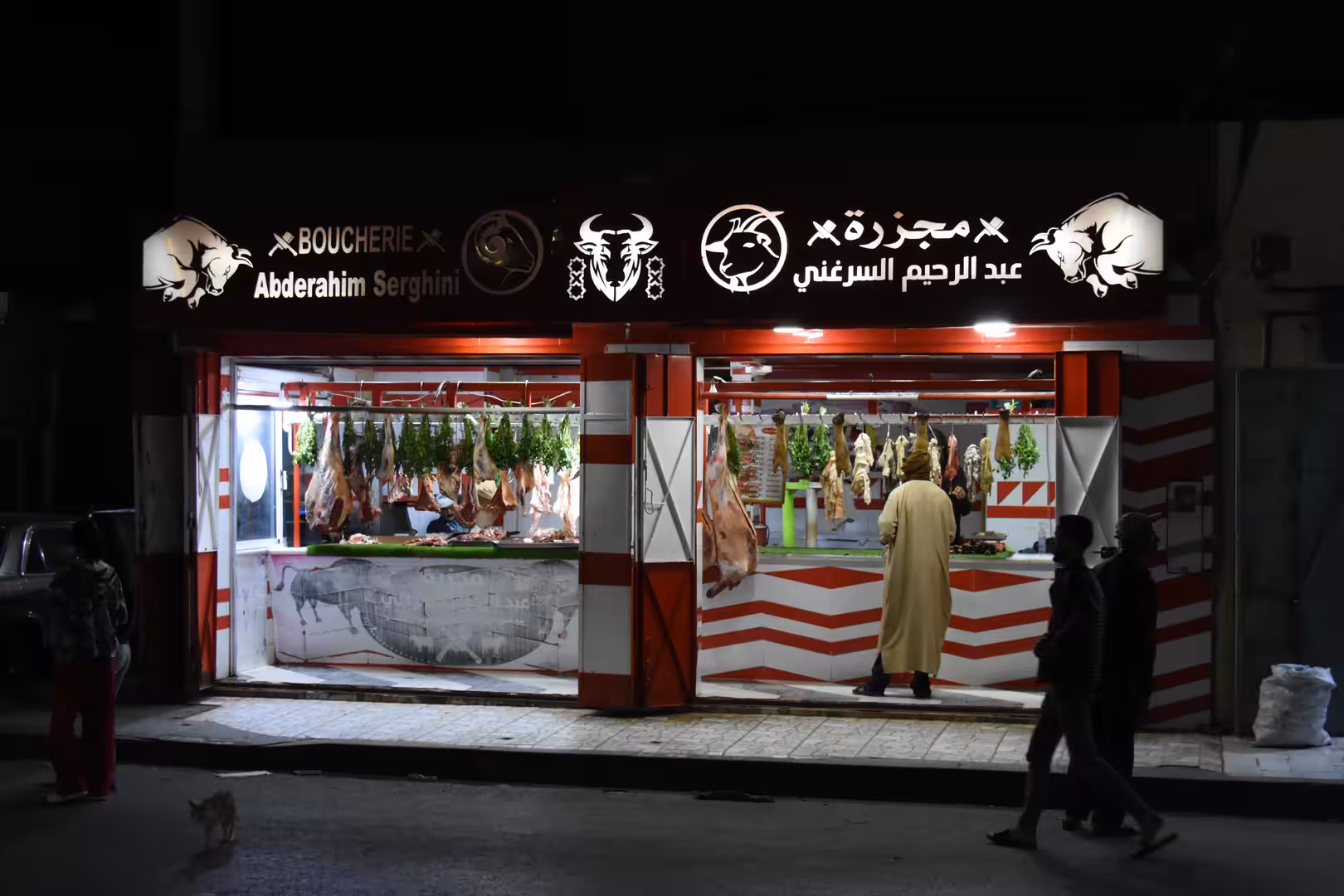 Illuminated butcher shop in Laâyoune at night with hanging cuts of meat visible through large open storefront windows and people passing outside.