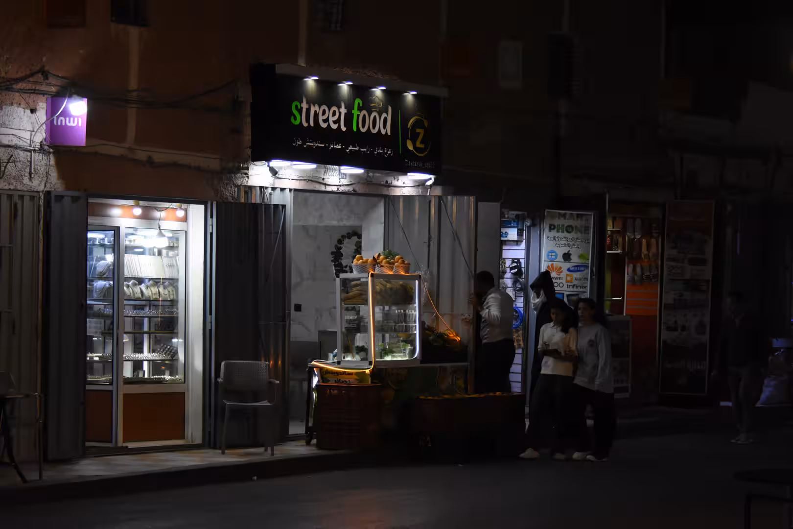 Nighttime street food storefront in Laâyoune with illuminated signage, a small food display and several people gathered outside.