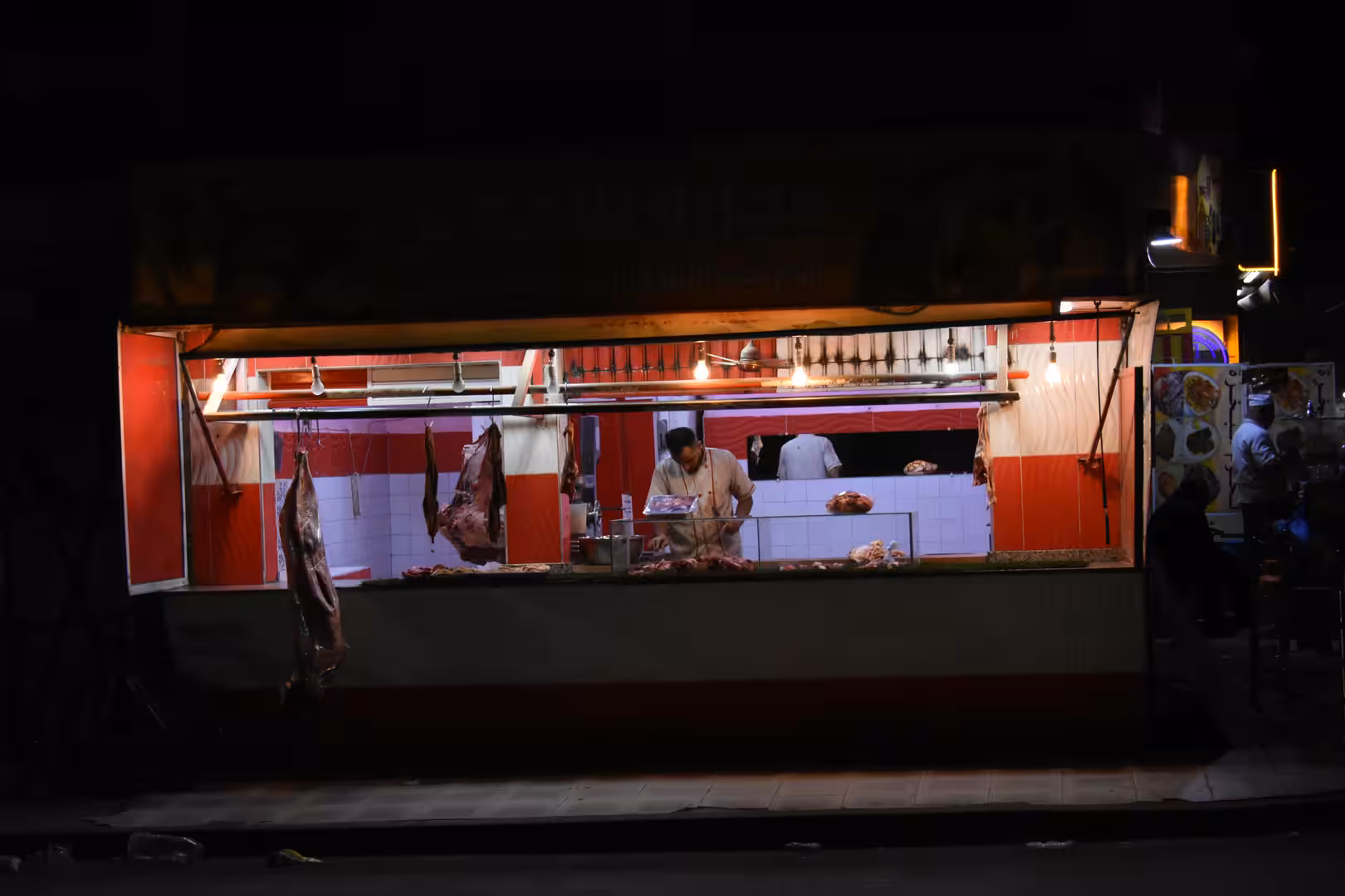 Nighttime view of an open butcher shop in Laâyoune with illuminated counters, hanging cuts of meat and staff preparing products inside.