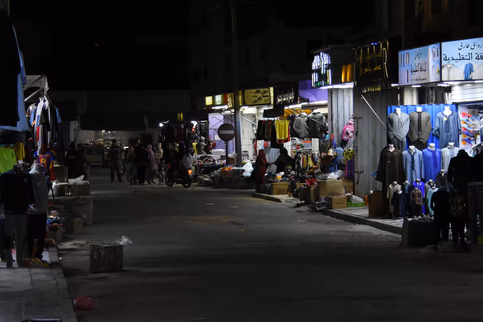 Evening street scene in Laâyoune showing illuminated clothing stores, garments displayed outside and people walking through a local shopping area.