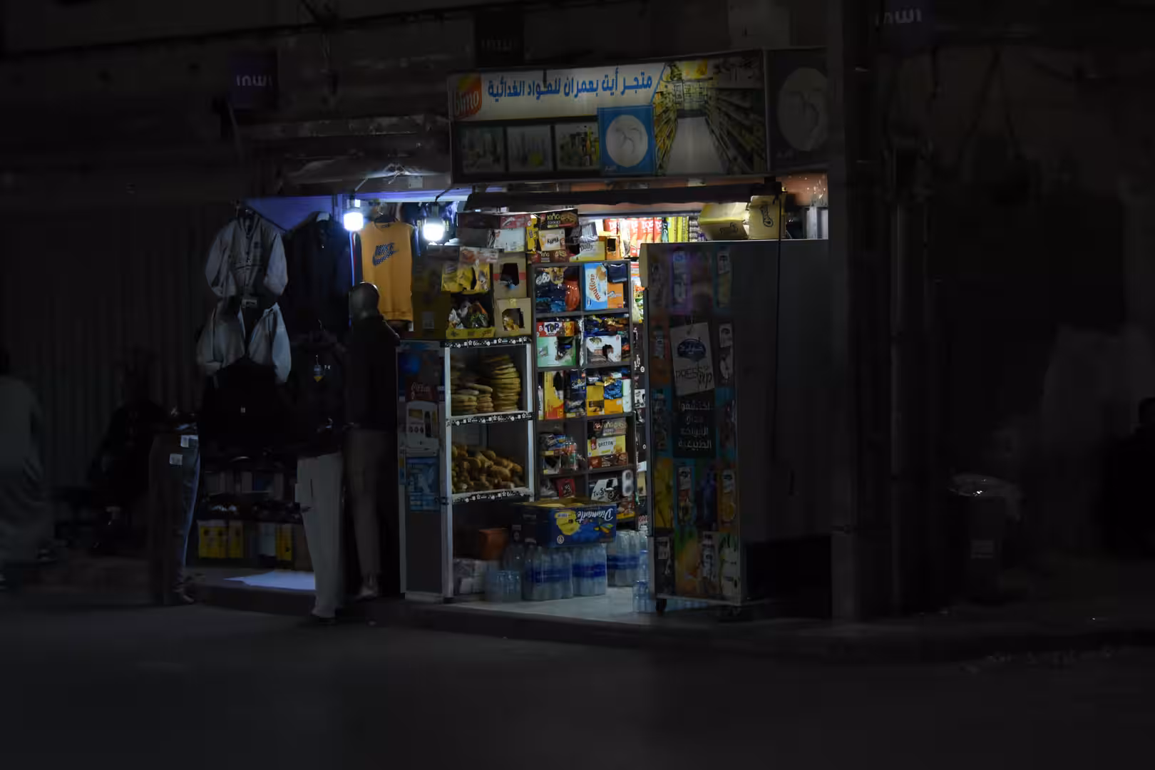 Small illuminated convenience store in Laâyoune at night with shelves of snacks, drinks, bottled water and a customer standing outside.