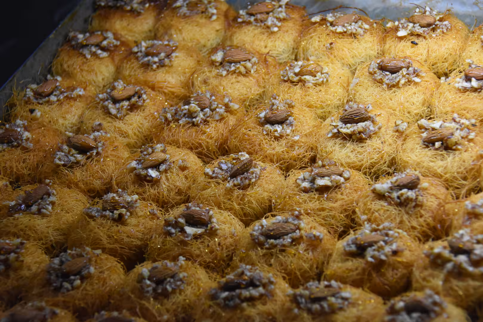 Close-up of golden shredded pastry nests topped with almonds and chopped nuts, displayed in a pastry tray.