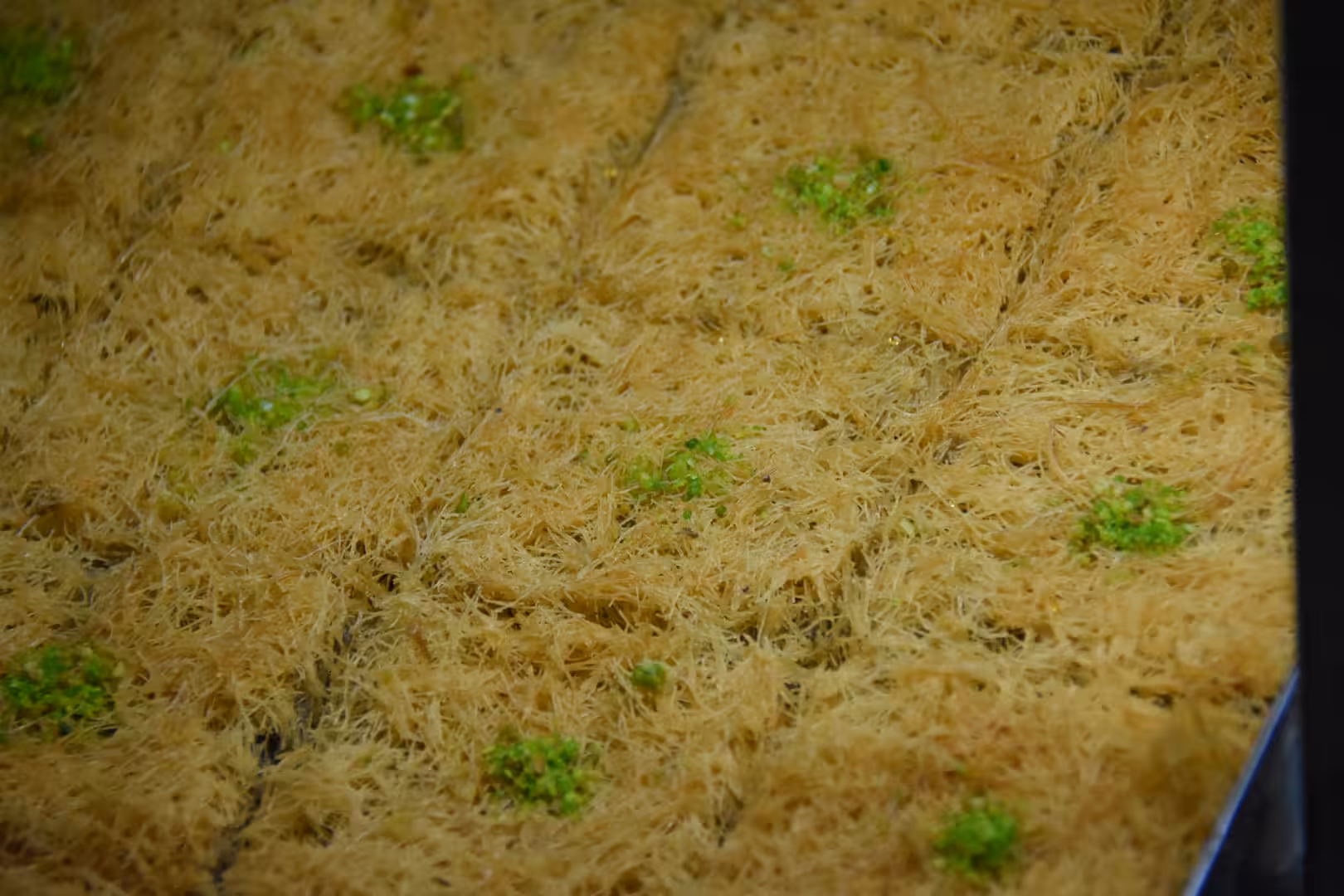 Close-up of golden fine shredded pastries arranged in a tray and topped with small portions of green pistachio garnish.