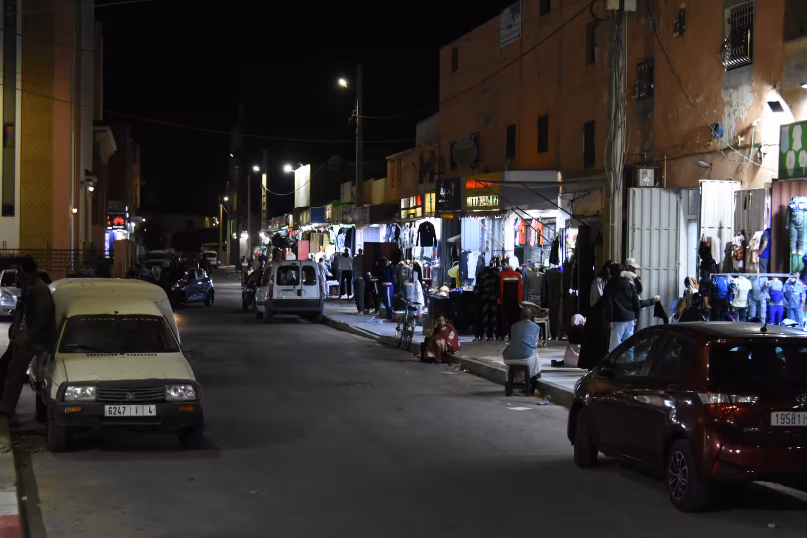 Nighttime street scene in Laâyoune with illuminated clothing shops, parked cars and people walking and shopping along the sidewalks.