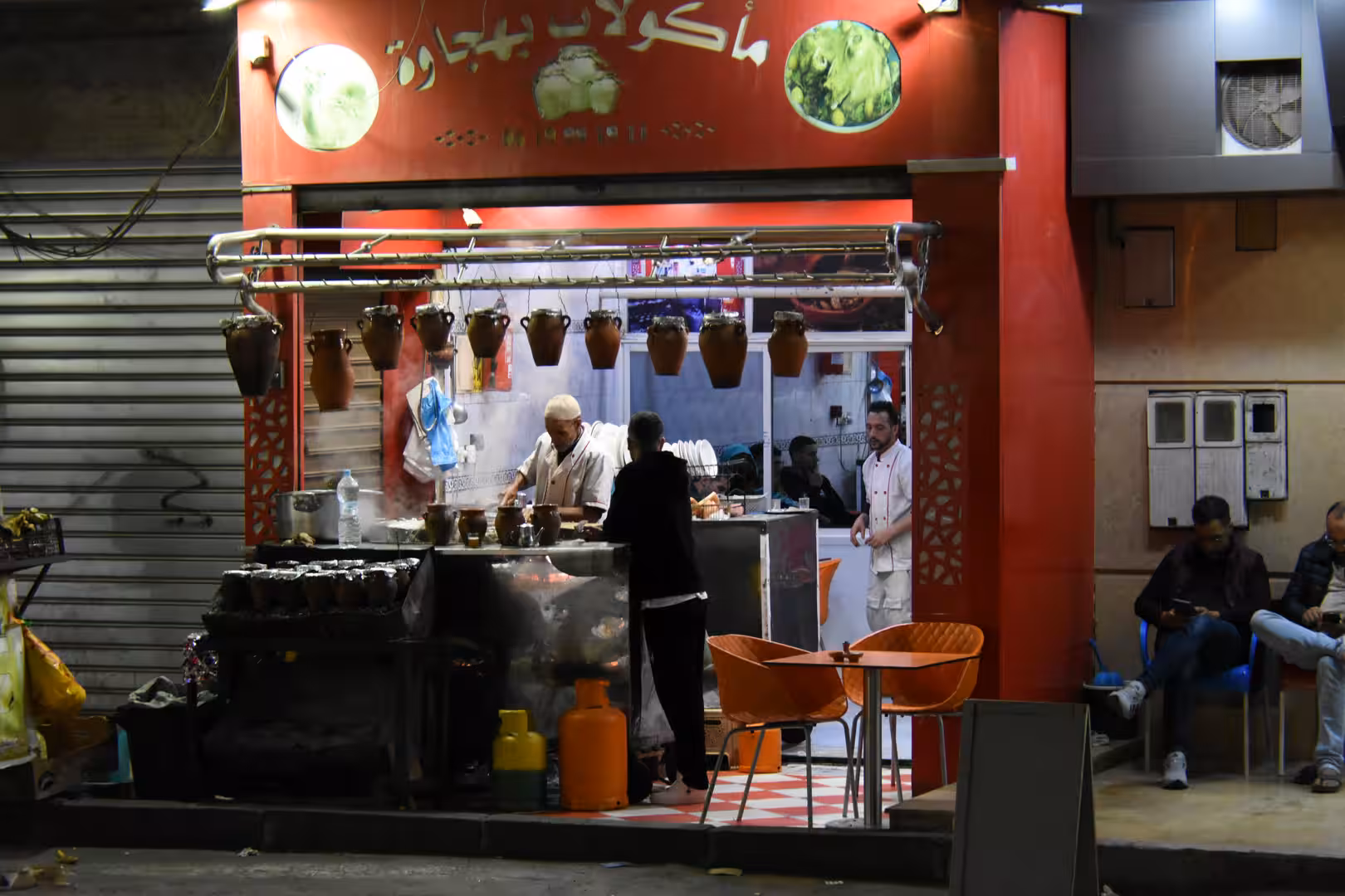 Nighttime street restaurant in Laâyoune with rows of hanging clay pots, cooks preparing food and customers seated outside.