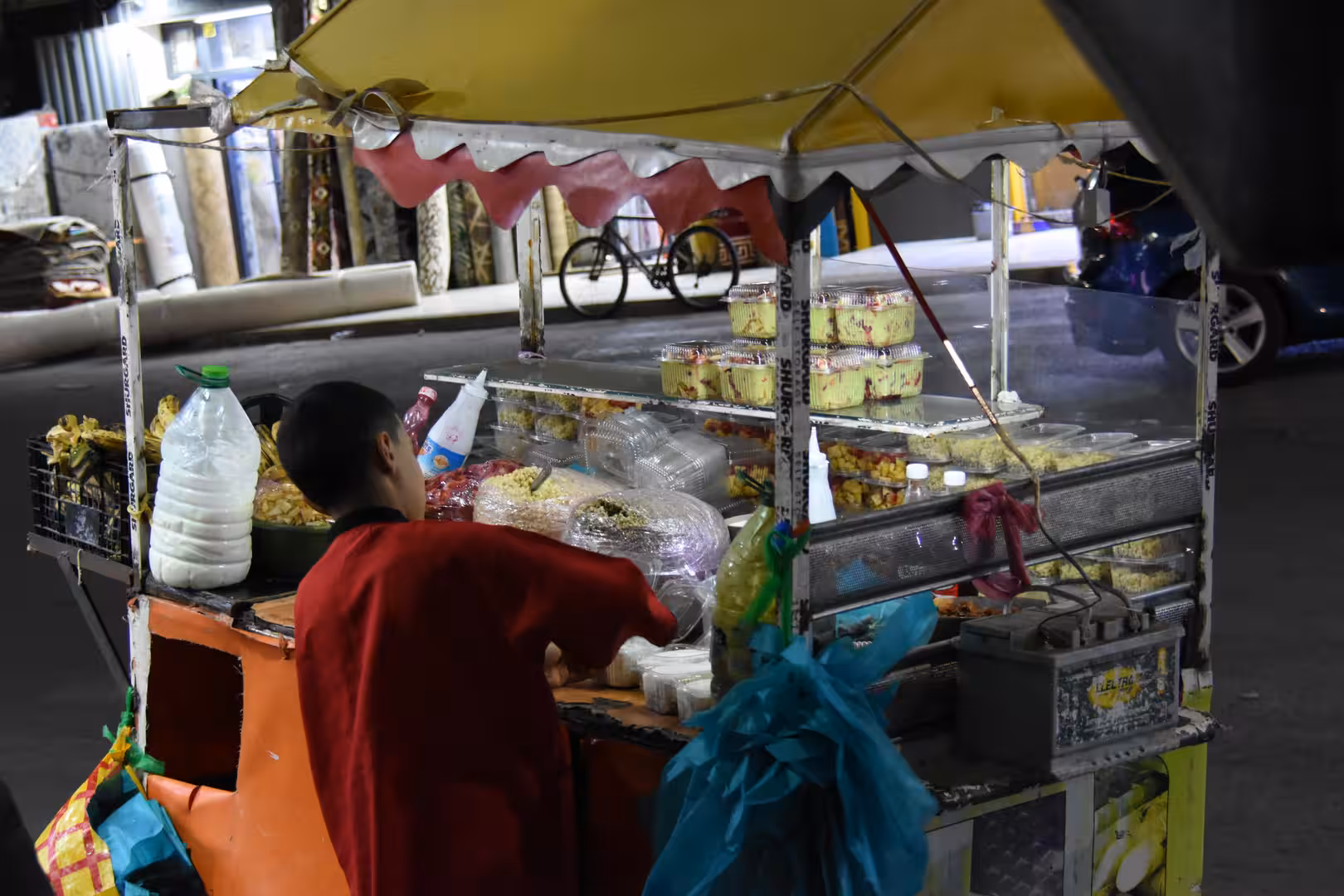 Nighttime street food cart in Laâyoune displaying prepared salads and packaged dishes while a vendor serves customers.