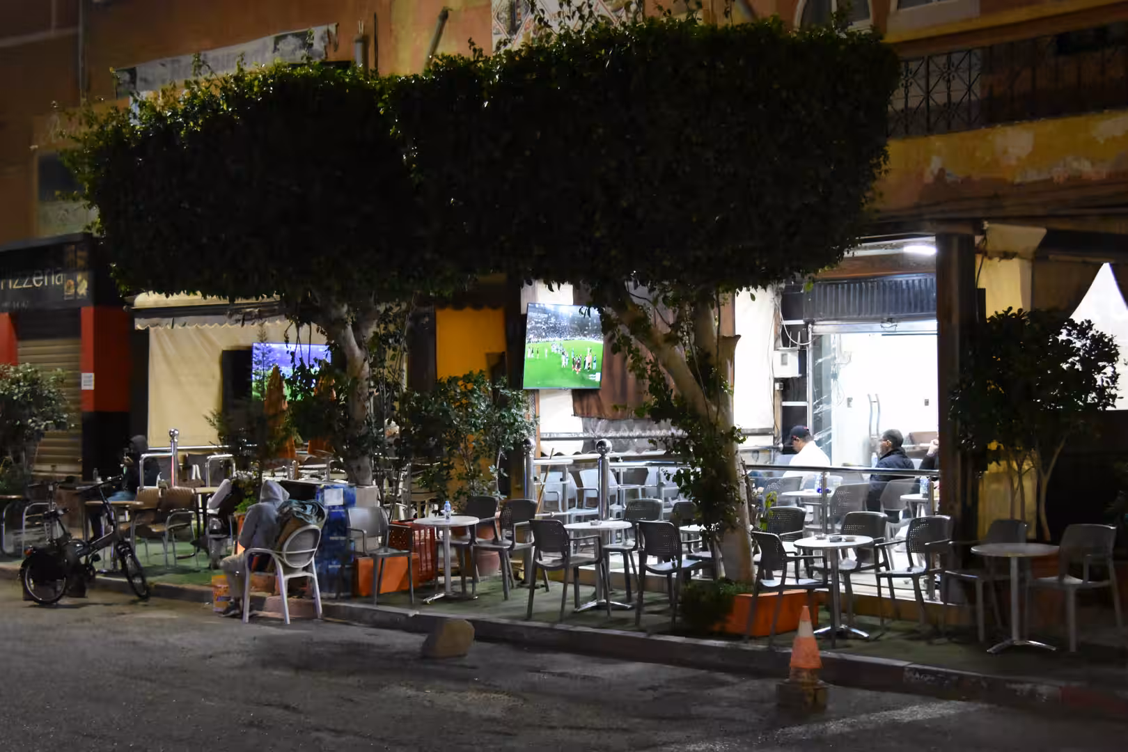 Outdoor café seating area in Laâyoune at night with tables beneath trimmed trees and people watching a football match.