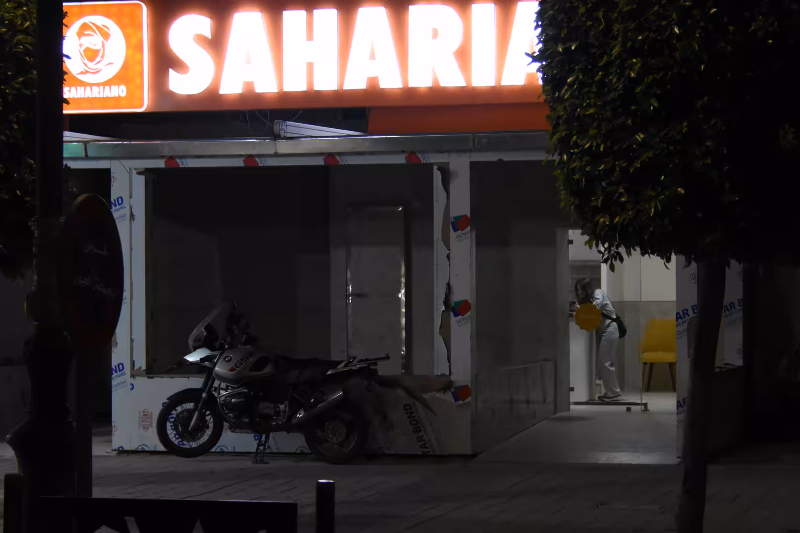 Night view of a newly fitted restaurant frontage in Laâyoune with illuminated Saharia sign, a parked motorcycle and a worker inside.