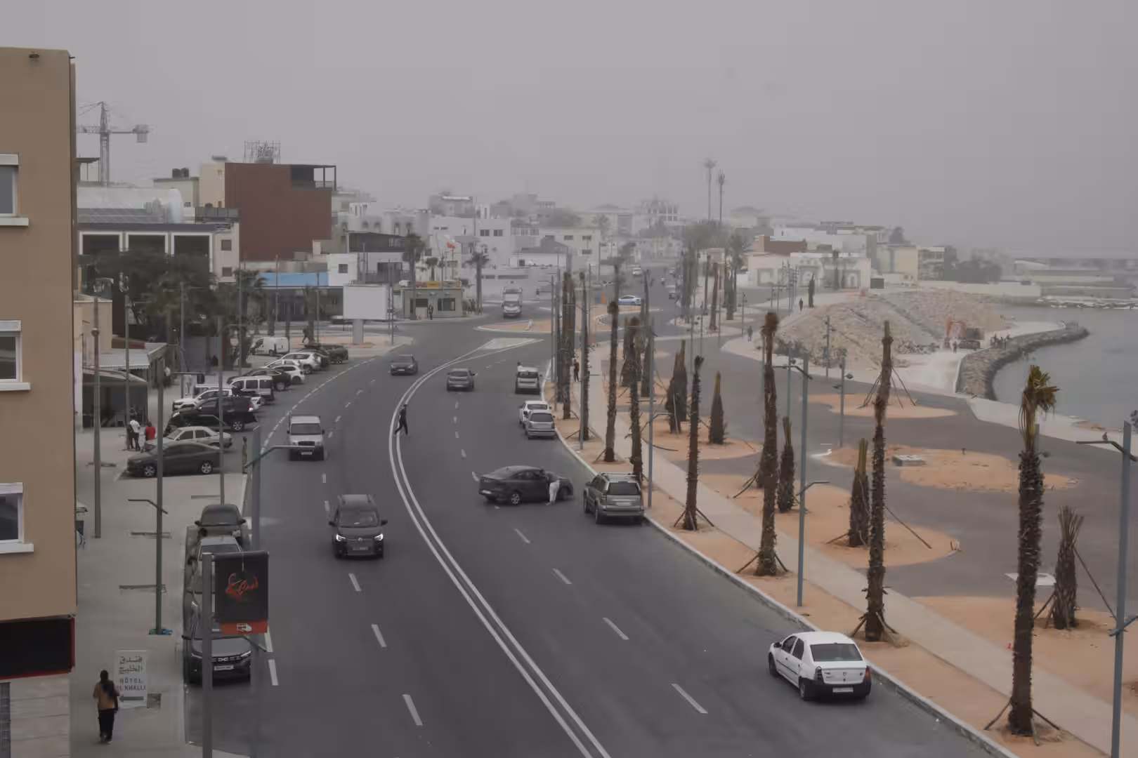 Elevated view of a wide coastal road with cars, palm trees and seaside development under a hazy sky.