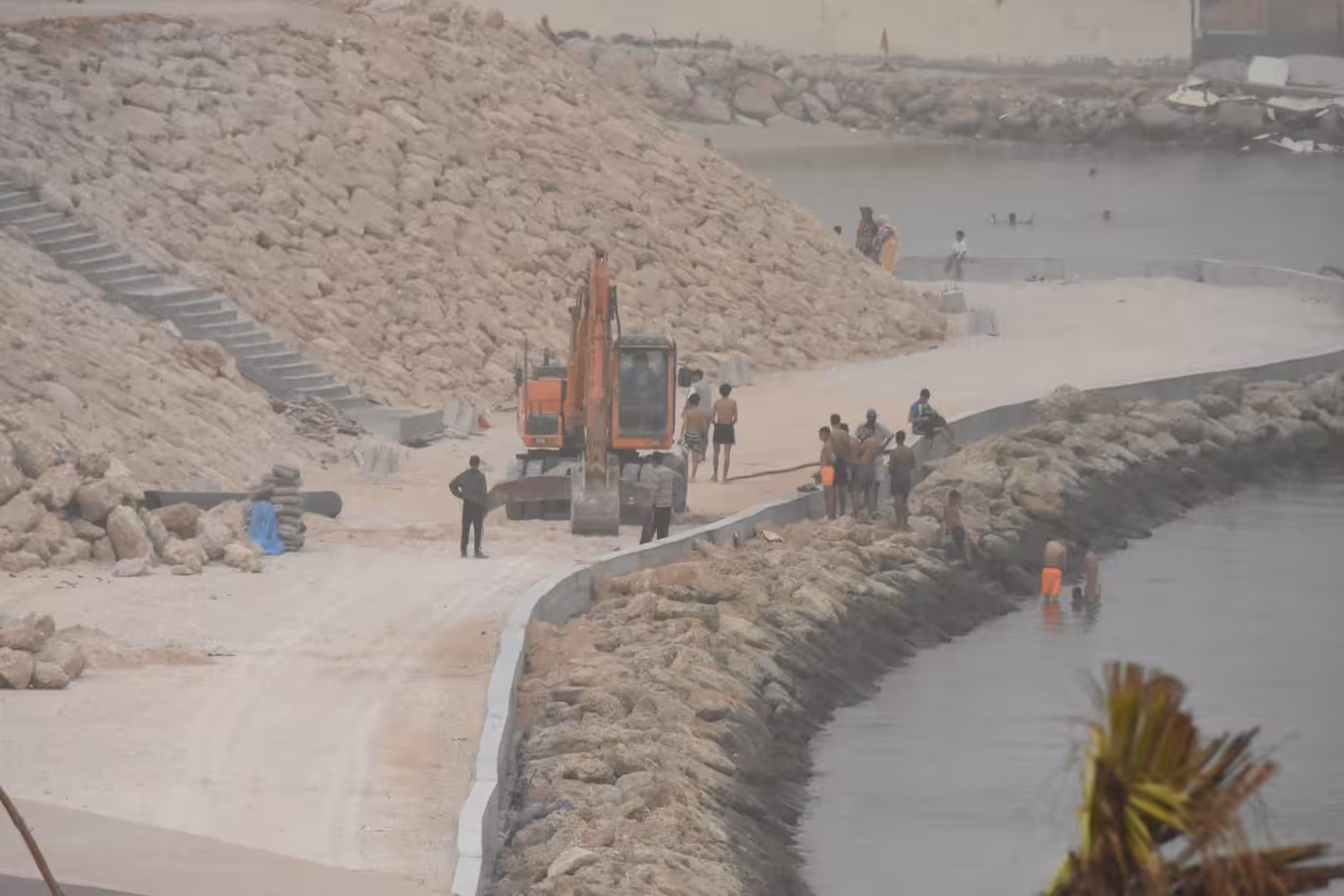 View of a coastal construction area with an excavator, stone embankment and people gathered near the water.