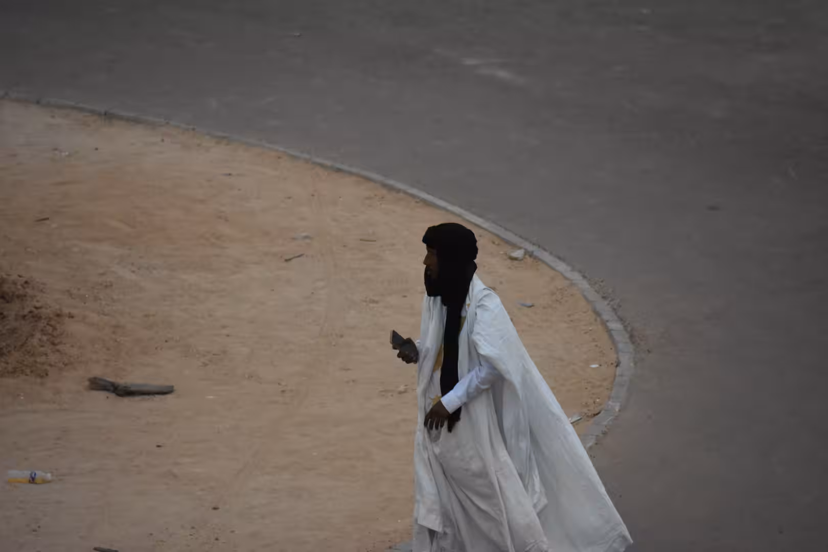 Person dressed in flowing white traditional clothing walking beside a curved road while holding a phone.