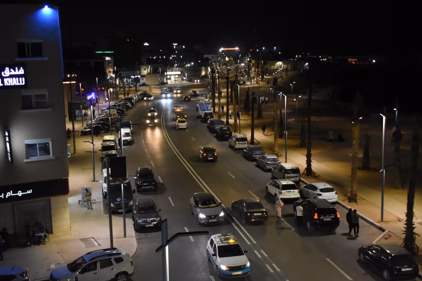 Elevated nighttime view of a busy boulevard with moving traffic, parked cars and illuminated sidewalks.
