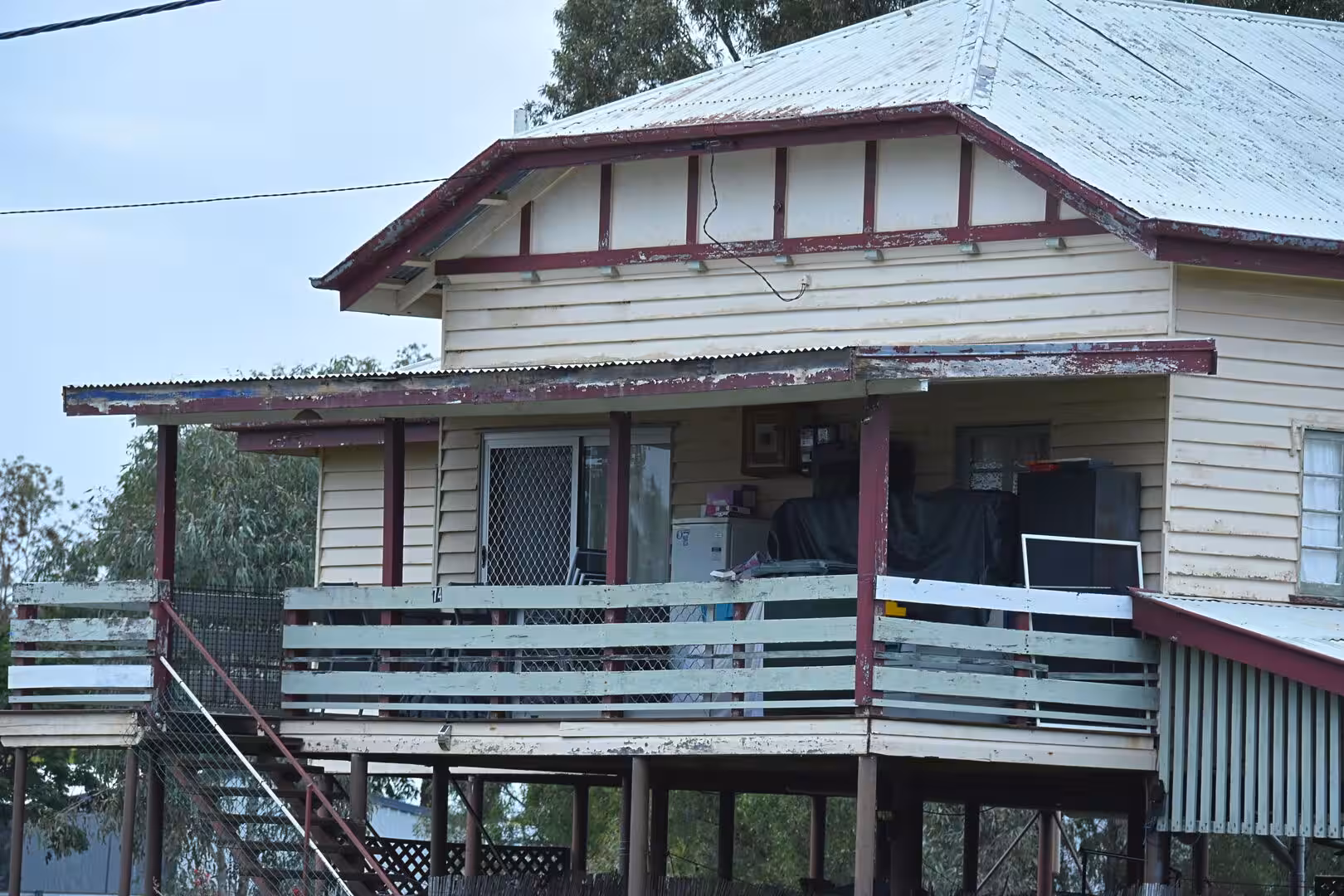 A weathered timber house on stilts has a wrap-around verandah crammed with stored items.