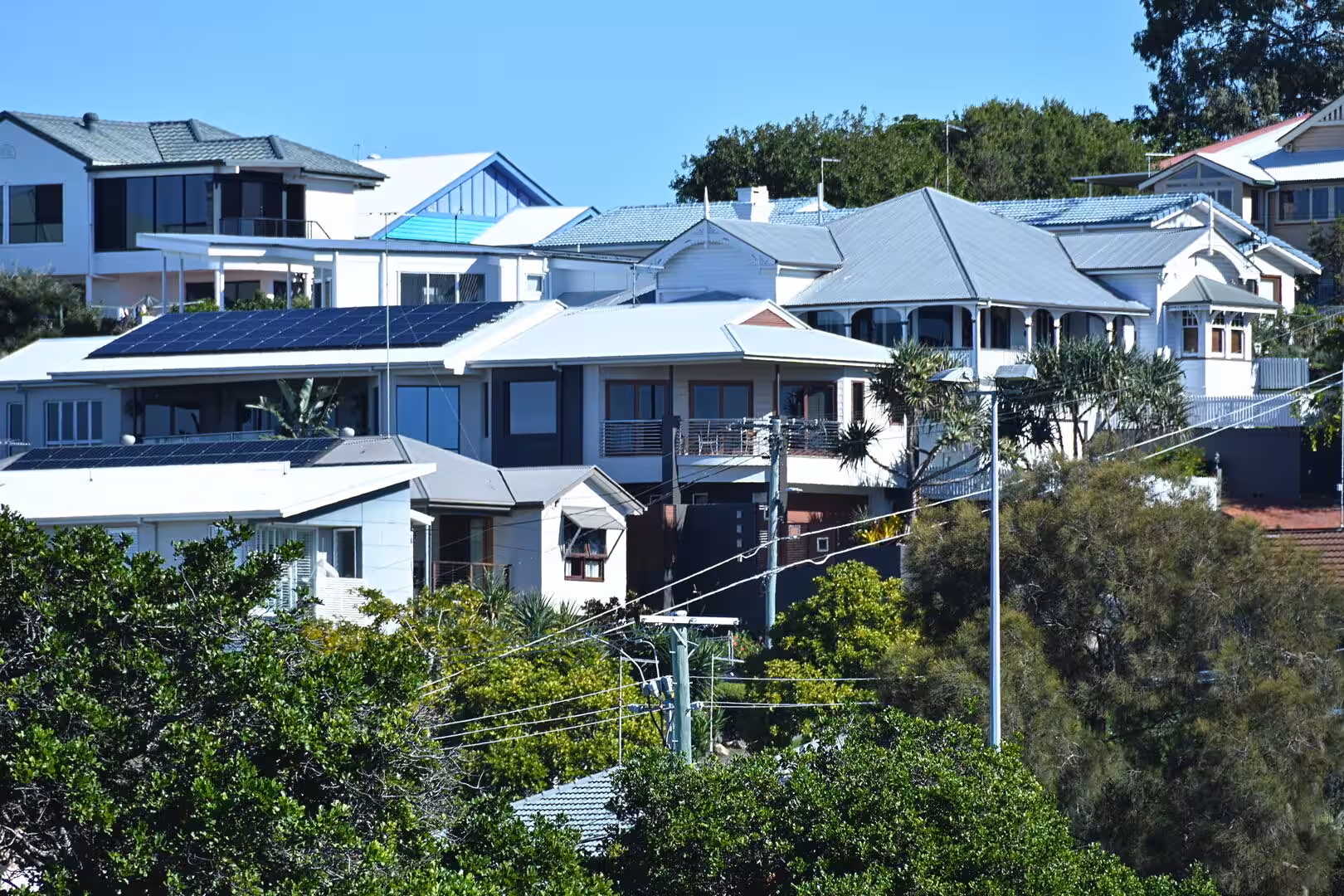Rows of white homes with rooftop solar panels sit on a leafy hill under blue sky.