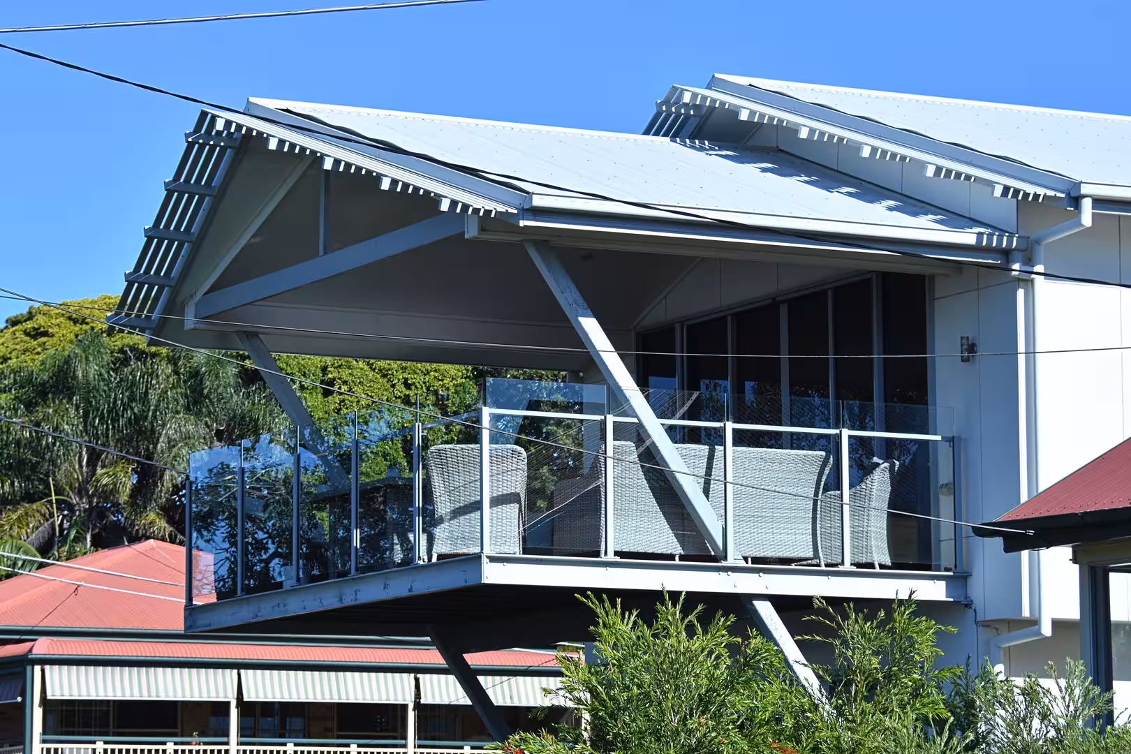 A modern two-storey home with an angular metal roof and a glass balcony lined with grey wicker chairs.