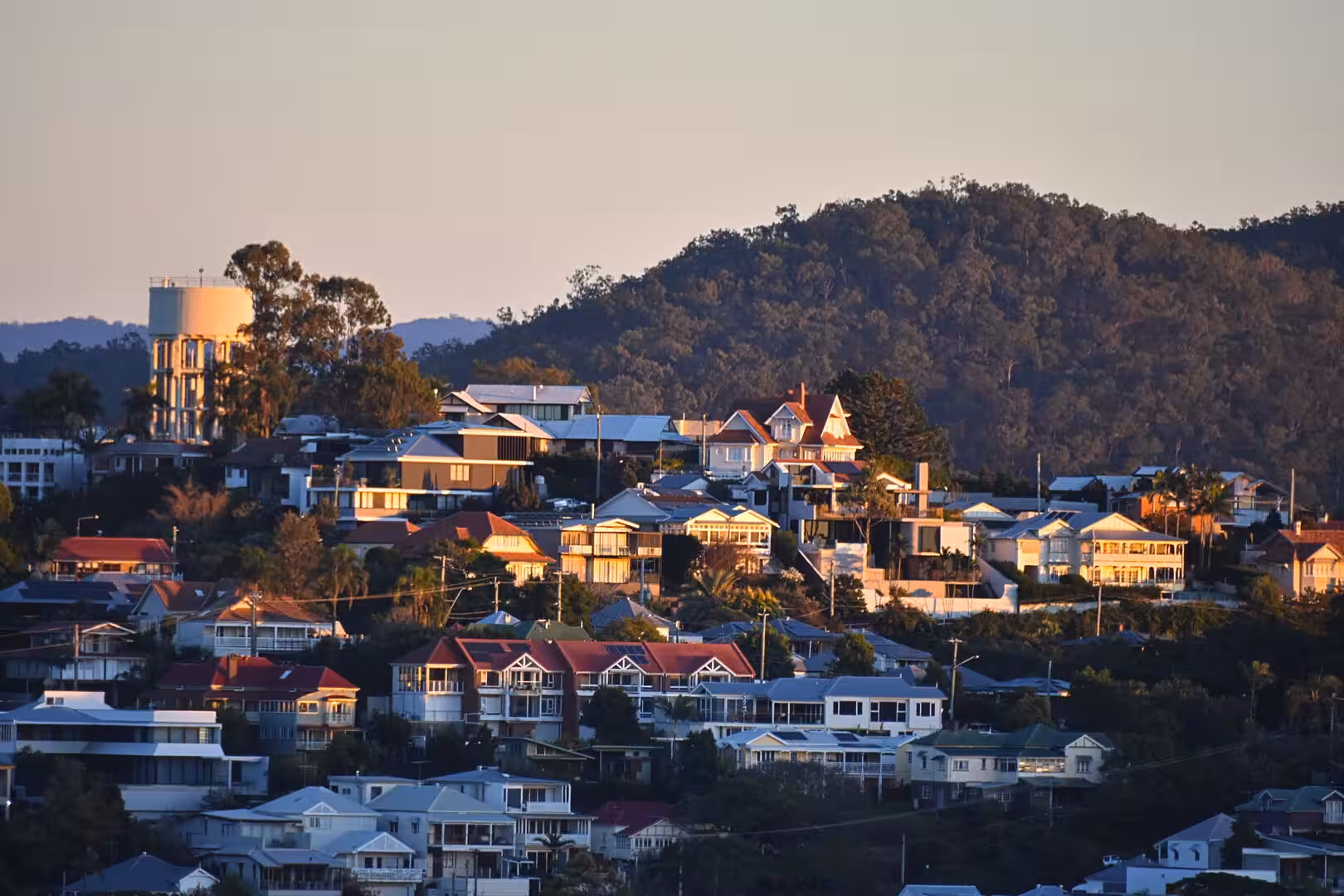 A sunset view over a hillside suburb with a white water tower and a forested ridge.