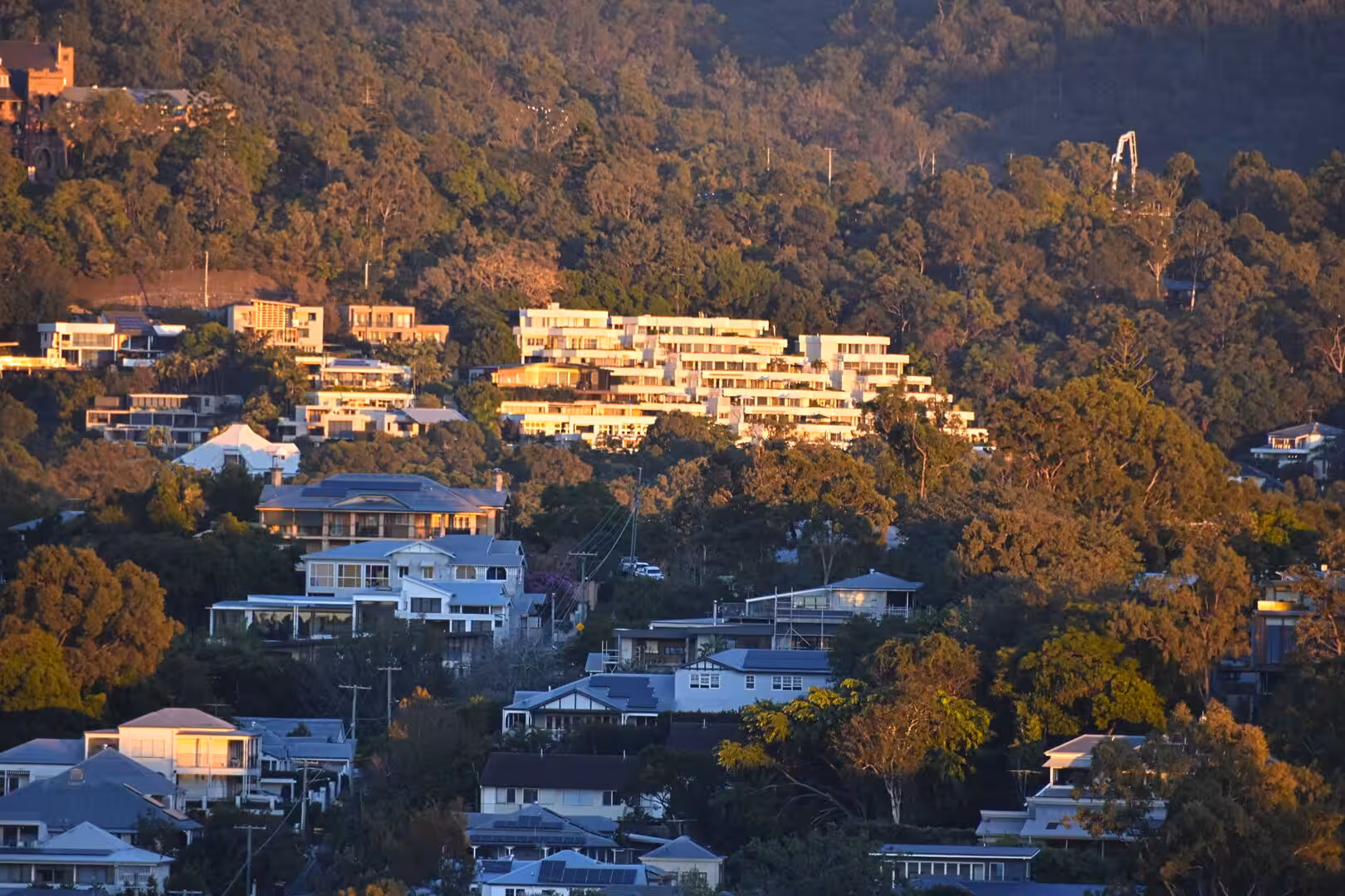 A long modern apartment complex glows amber on a wooded hillside above scattered homes.