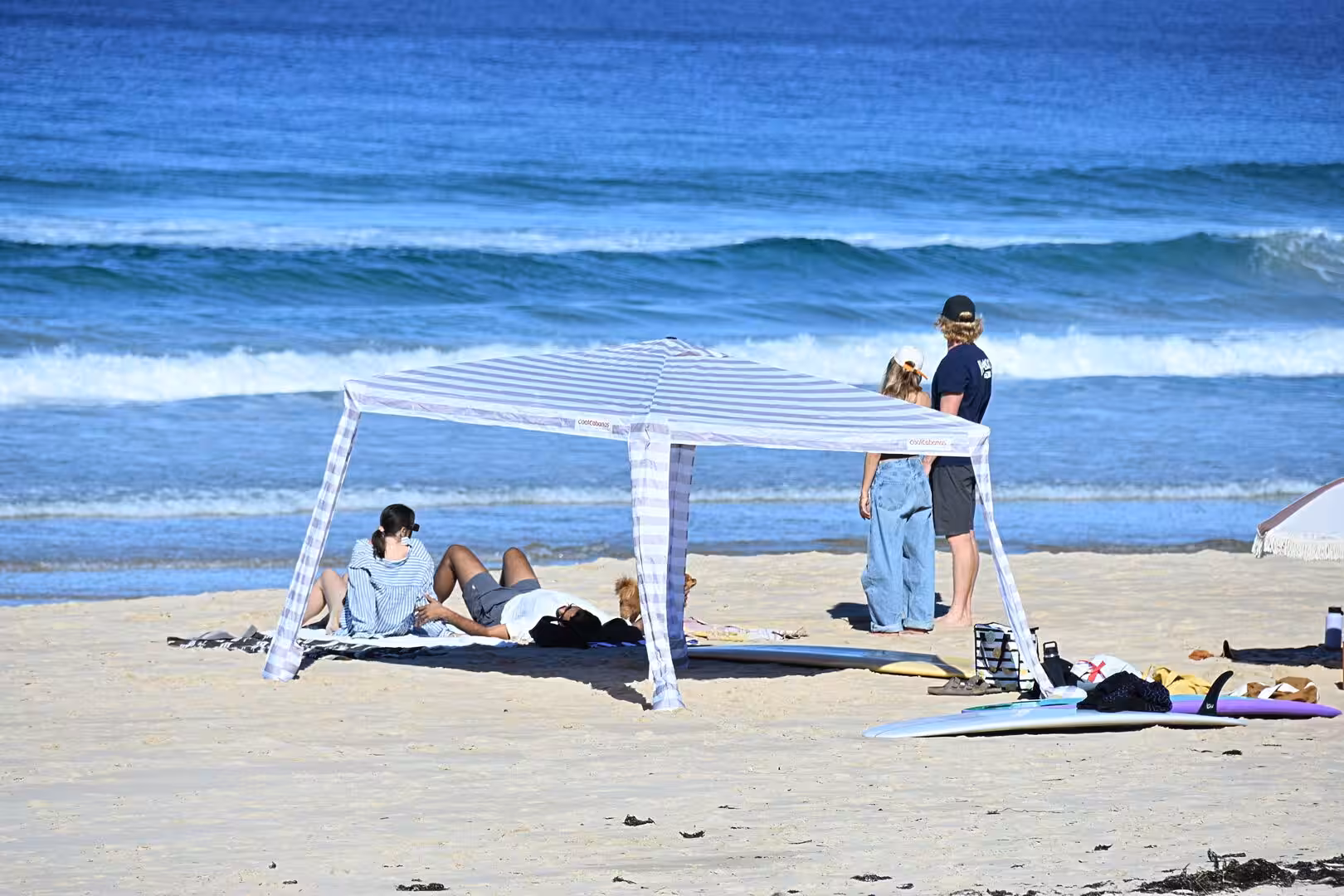 People lounge under a striped canvas tent on the sand by the surf.