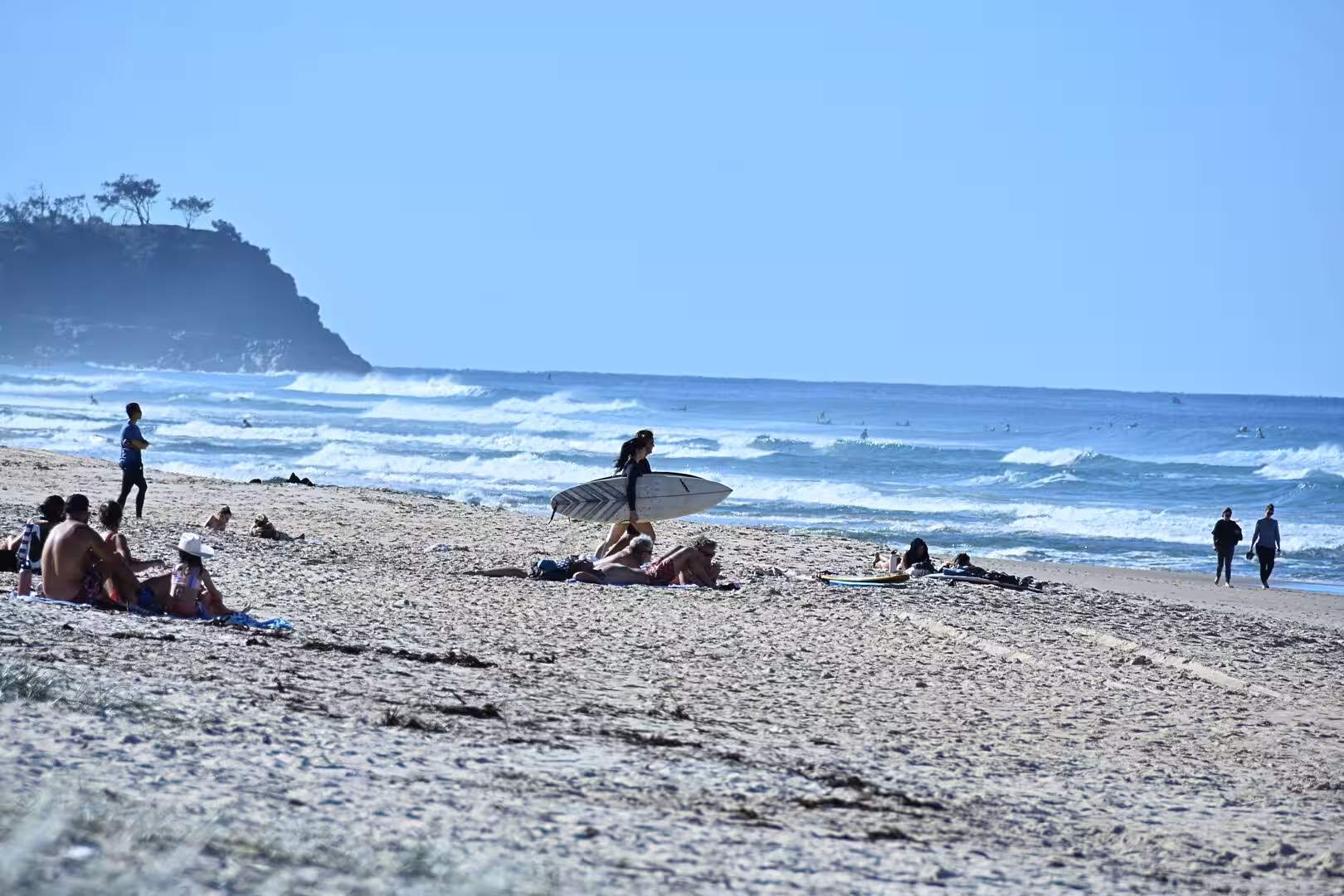A woman walks across a sandy beach carrying a surfboard beside a tree-topped headland.