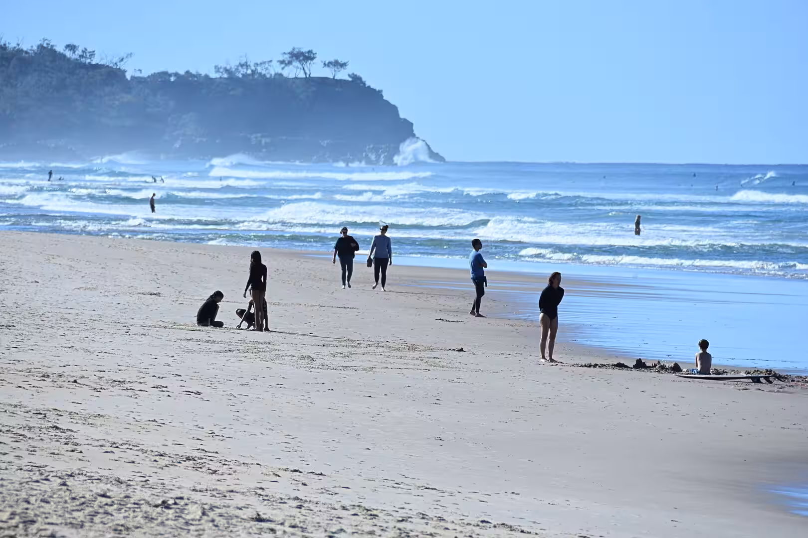 People walk and sit on a wide sandy beach as waves break beside a distant wooded headland.