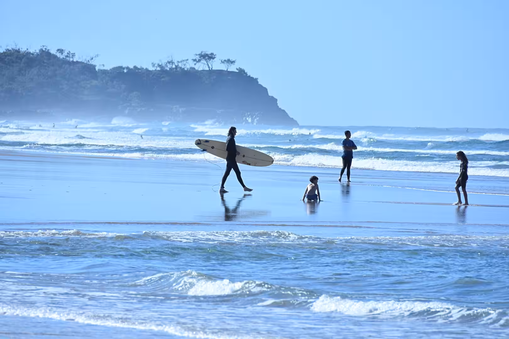 A surfer walks across glistening wet sand carrying a board toward the breaking waves.
