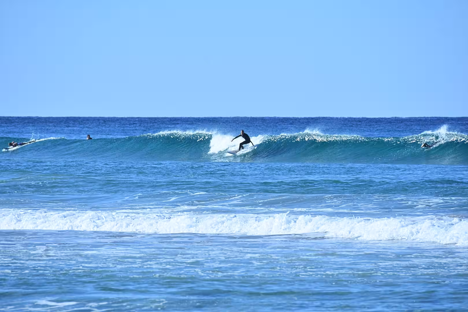 A surfer rides a curling turquoise wave with white spray as the blue sea stretches to the horizon.