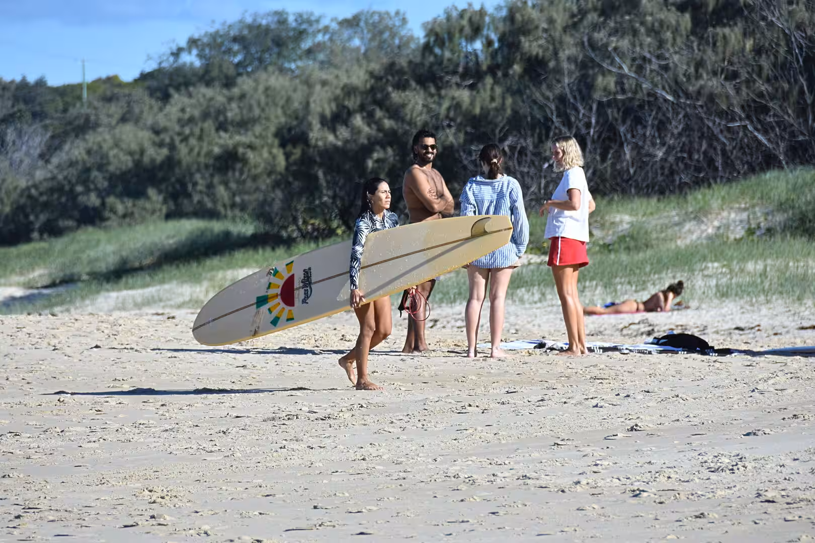 A young woman holds a surfboard while a group of friends stand together on the sand.